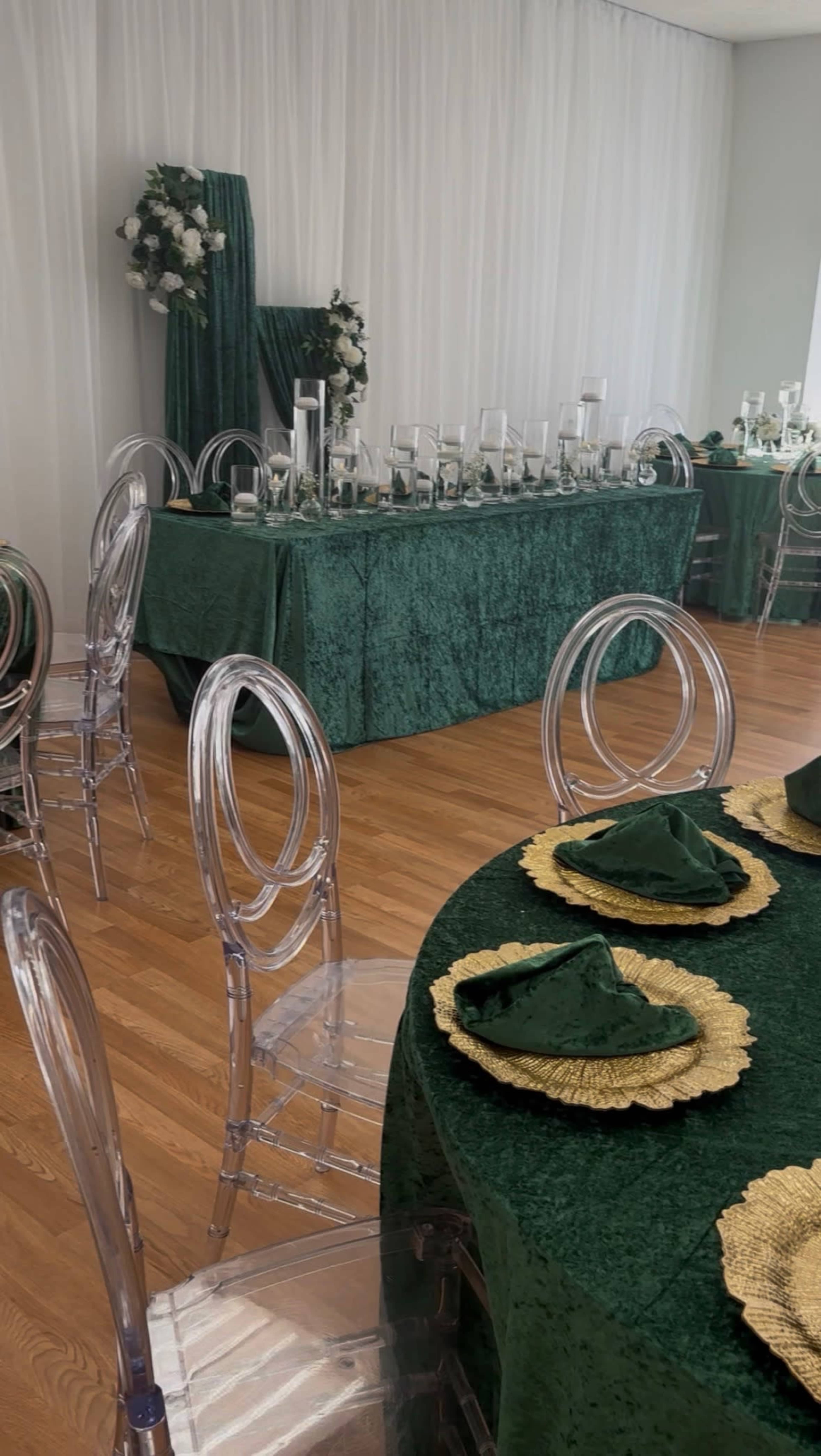 The image shows a beautifully arranged banquet hall with green textured tablecloths, clear acrylic chairs, and a long table set with glass decor and floral accents.