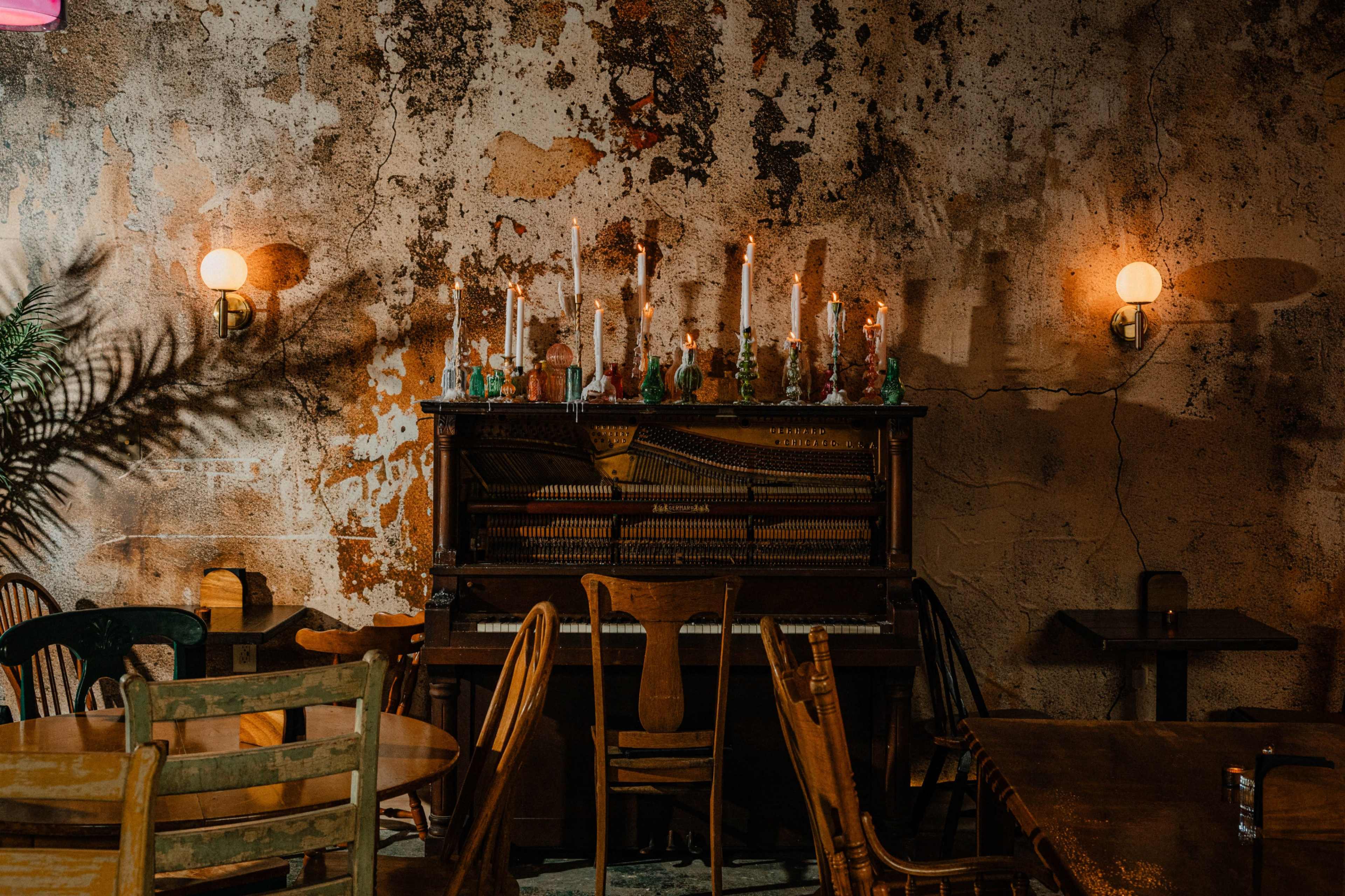 The image shows an old piano with candles arranged on top, set against a textured wall in a dimly lit, rustic café with wooden furniture.