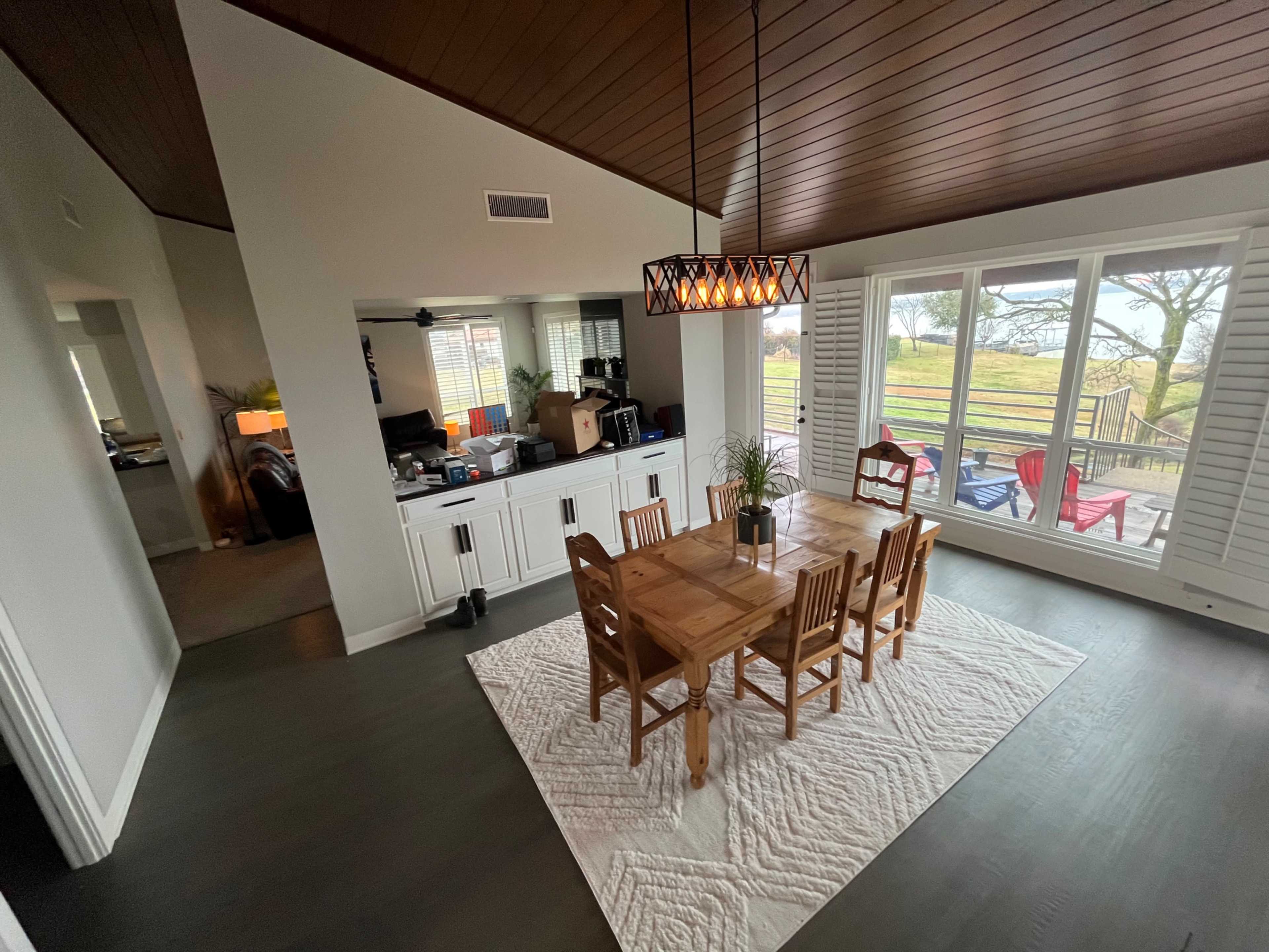 The image shows a dining area with a wooden table surrounded by chairs, overlooking a large window that reveals a view of a grassy landscape and colorful outdoor chairs.