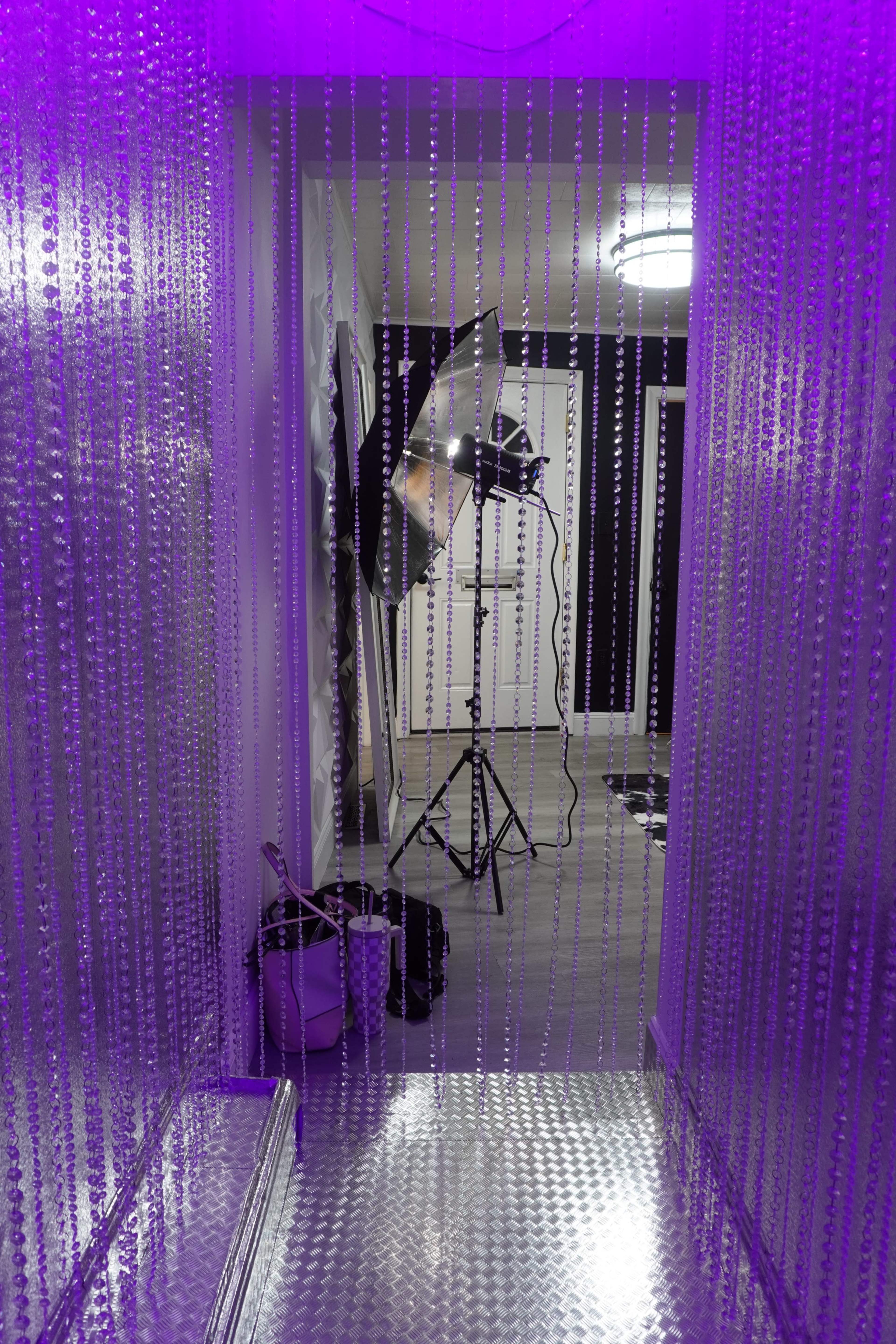 A hallway lined with beaded curtains leads to a photography setup with a light and tripod in the background.