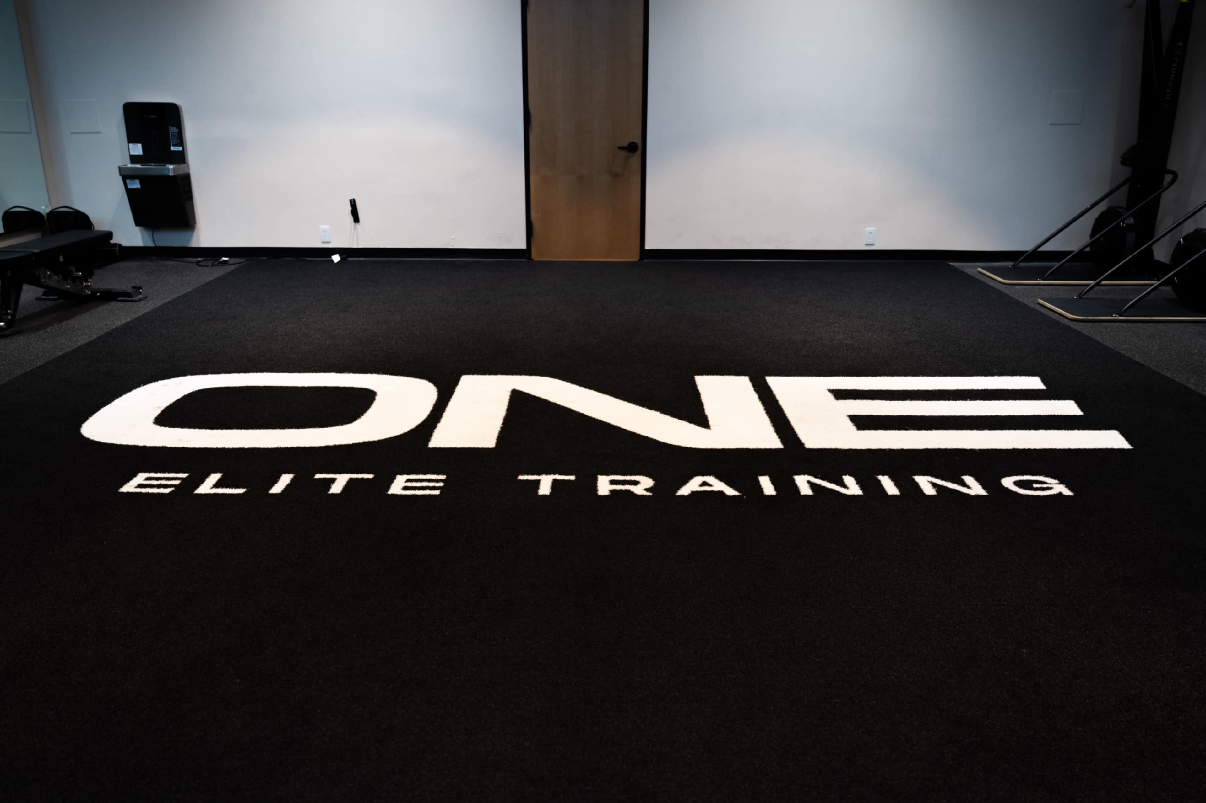 The image shows a fitness training area with the words "ONE ELITE TRAINING" prominently displayed on the black carpet.