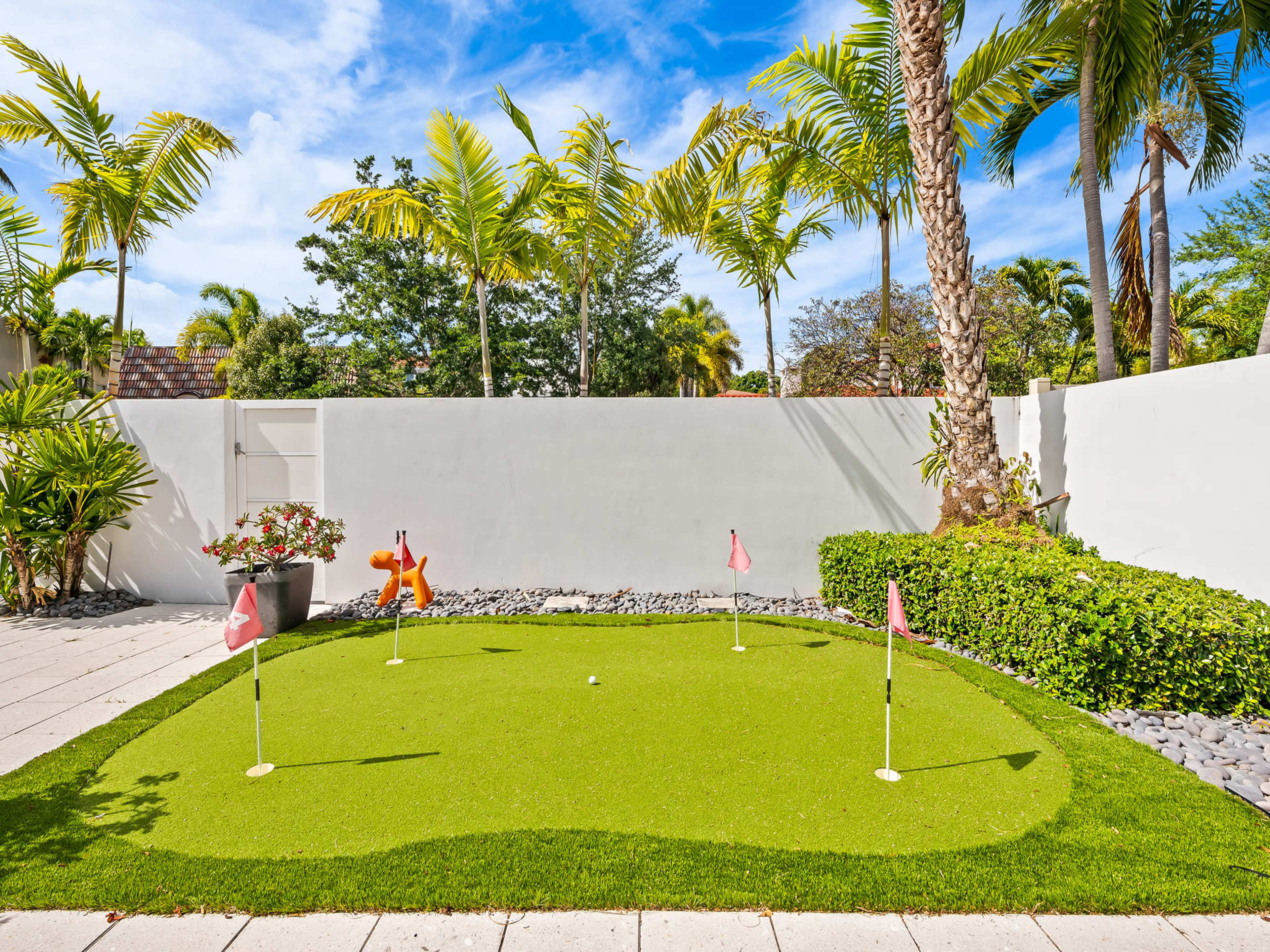 The image shows a small outdoor putting green surrounded by tropical plants and trees, with a white wall in the background.