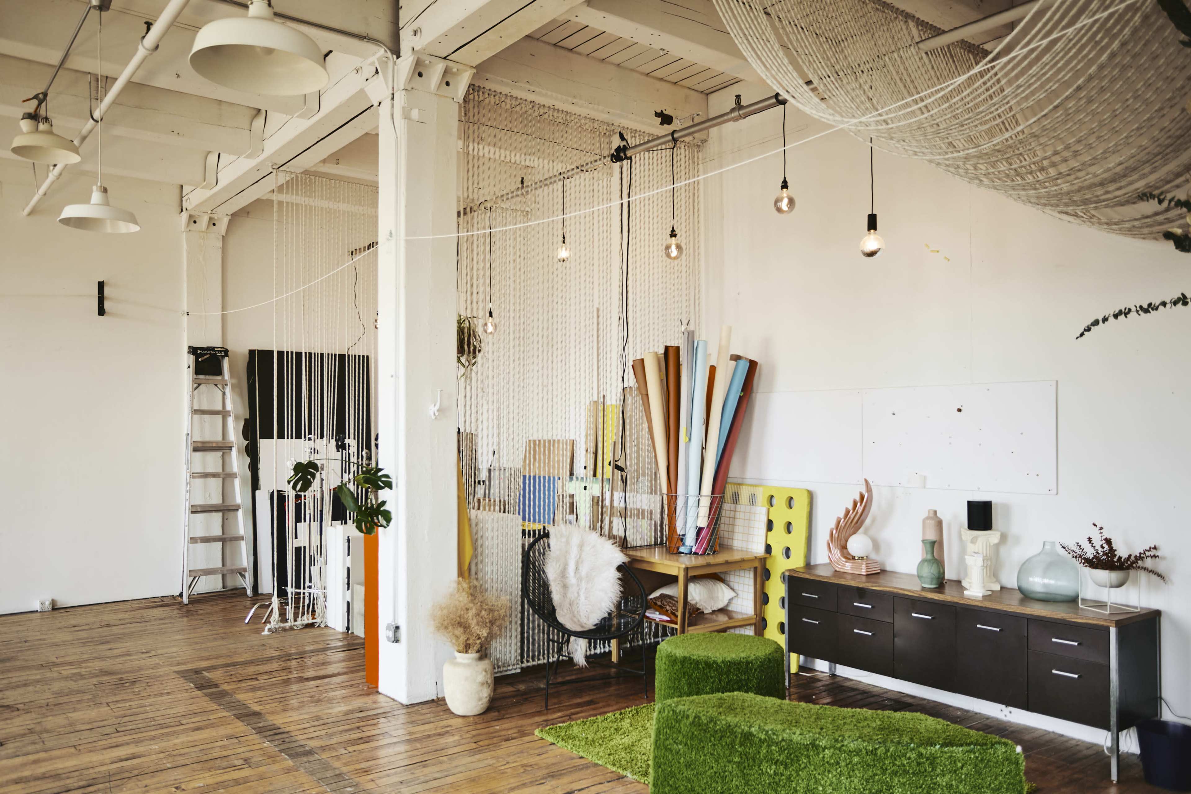 An interior space features a variety of colorful artistic materials, a ladder, and decorative elements against a white wall with wooden flooring.
