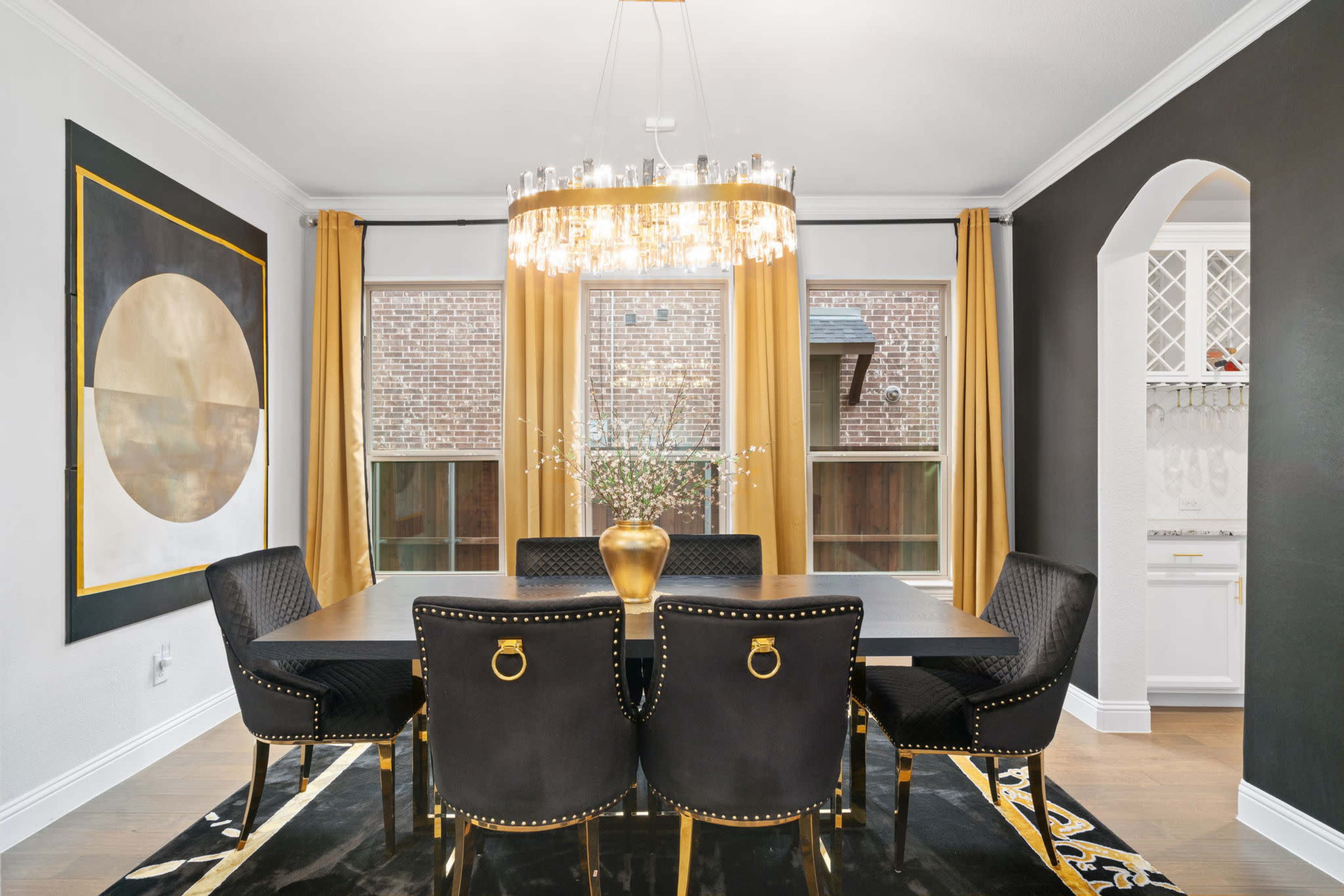 A modern dining room features a large table surrounded by black chairs, a chandelier overhead, and large windows dressed with golden curtains.