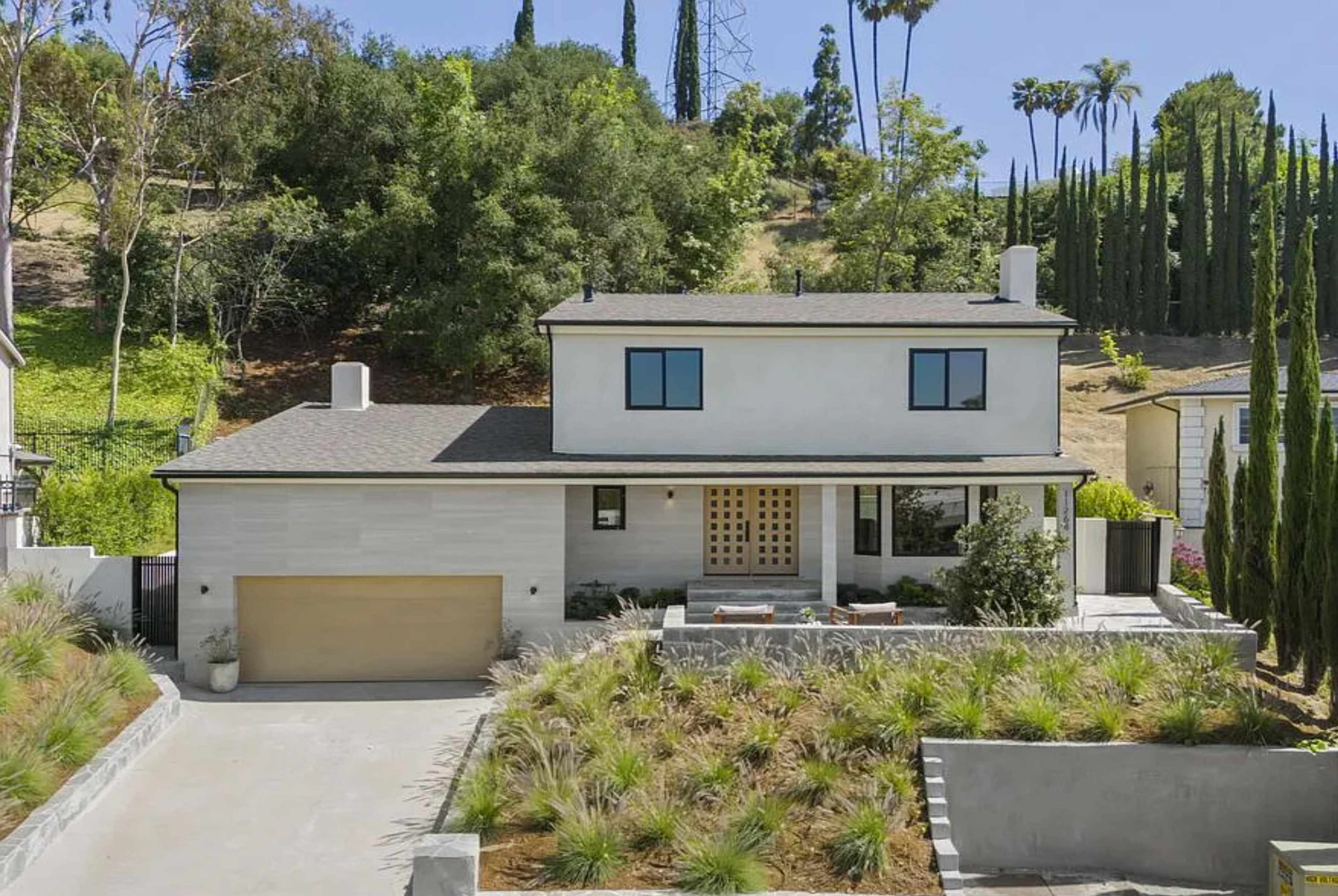 A two-story modern house with a landscaped front yard and a driveway sits against a backdrop of trees and hills.