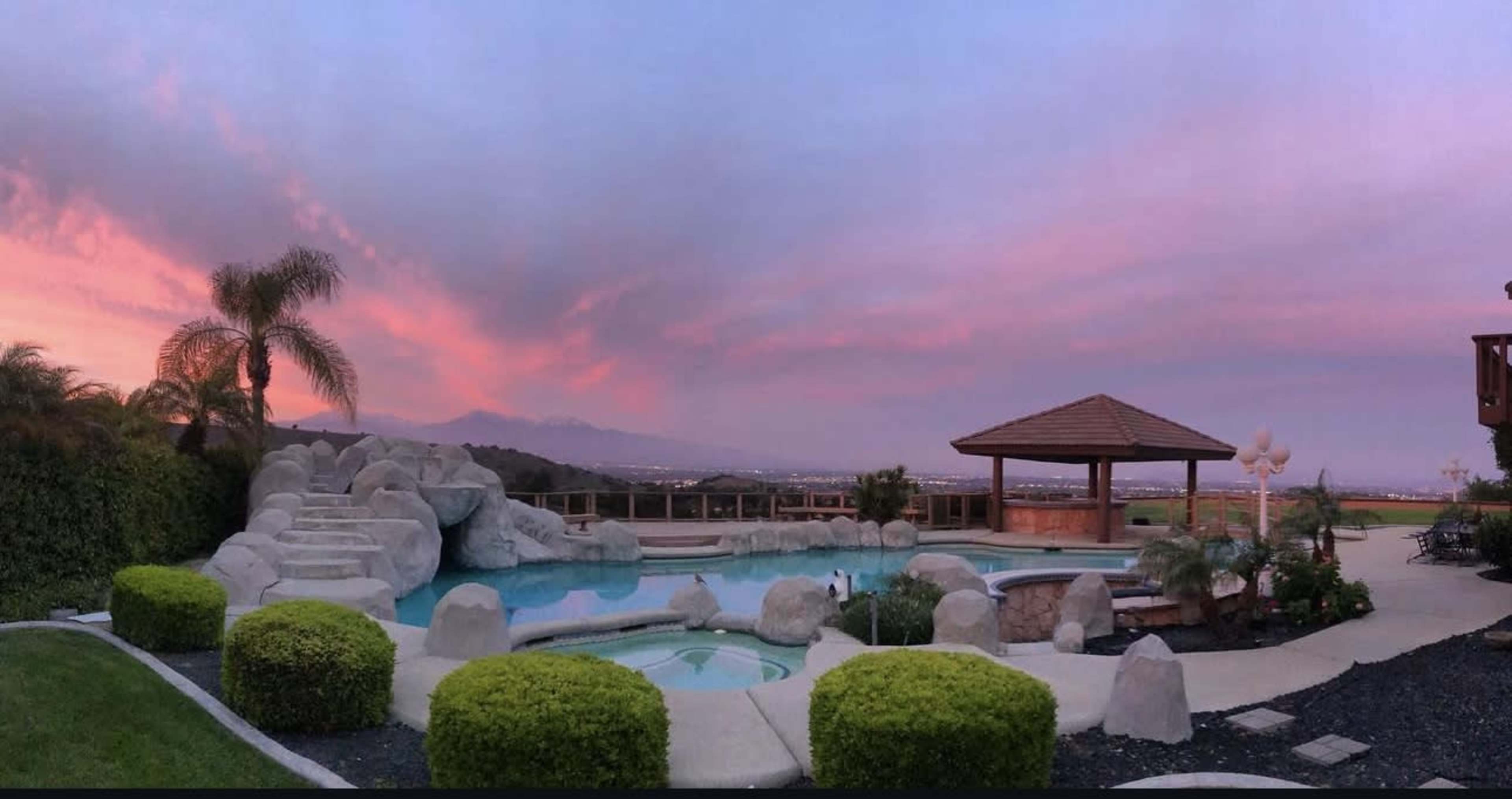 The image shows a backyard pool area with rock formations, a gazebo, and lush landscaping under a colorful sunset sky.