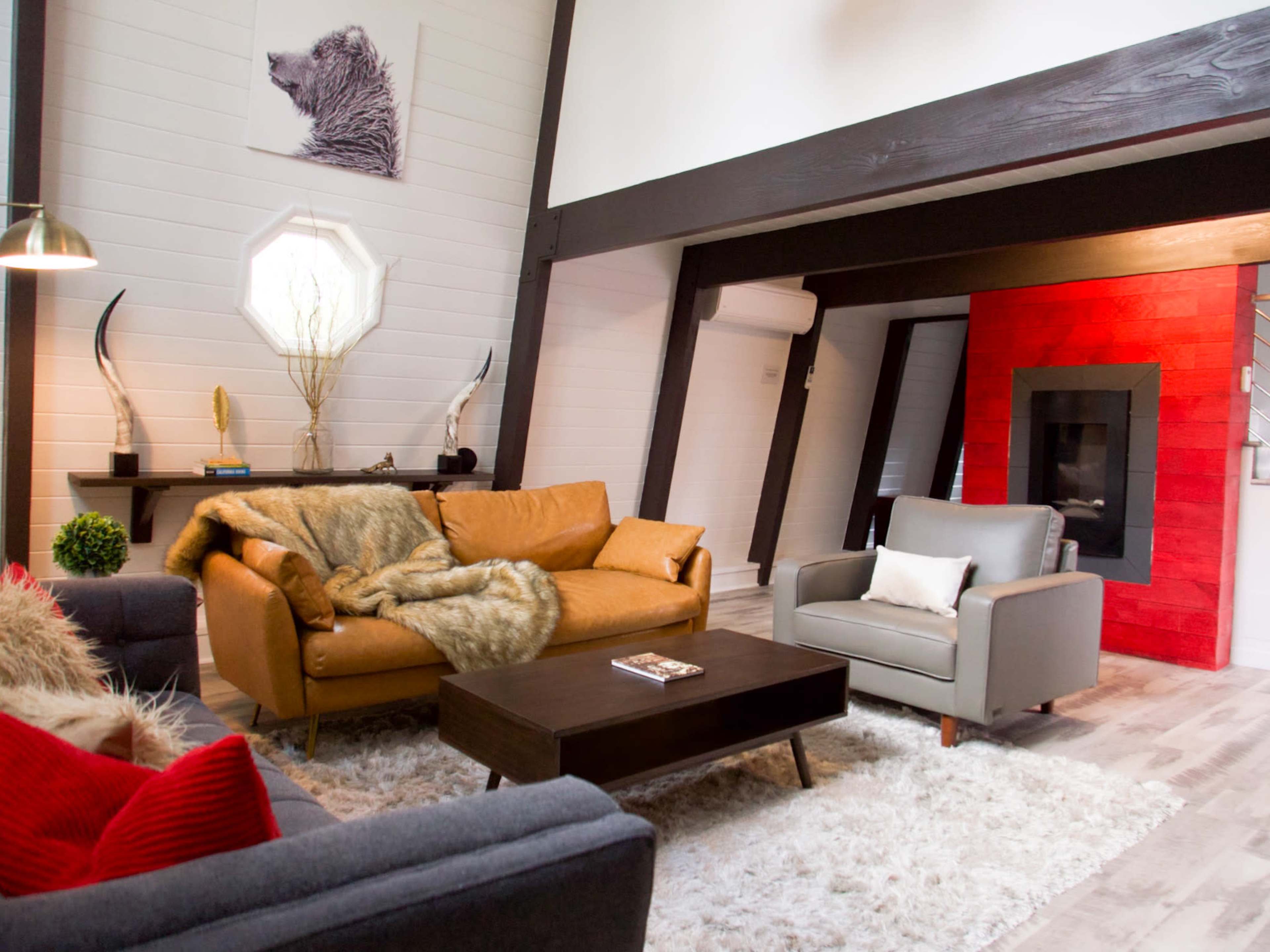 A modern living room with a mix of furniture, including a brown leather sofa, a gray armchair, and a coffee table, set against a backdrop of wooden beams and a red accent wall with a fireplace.