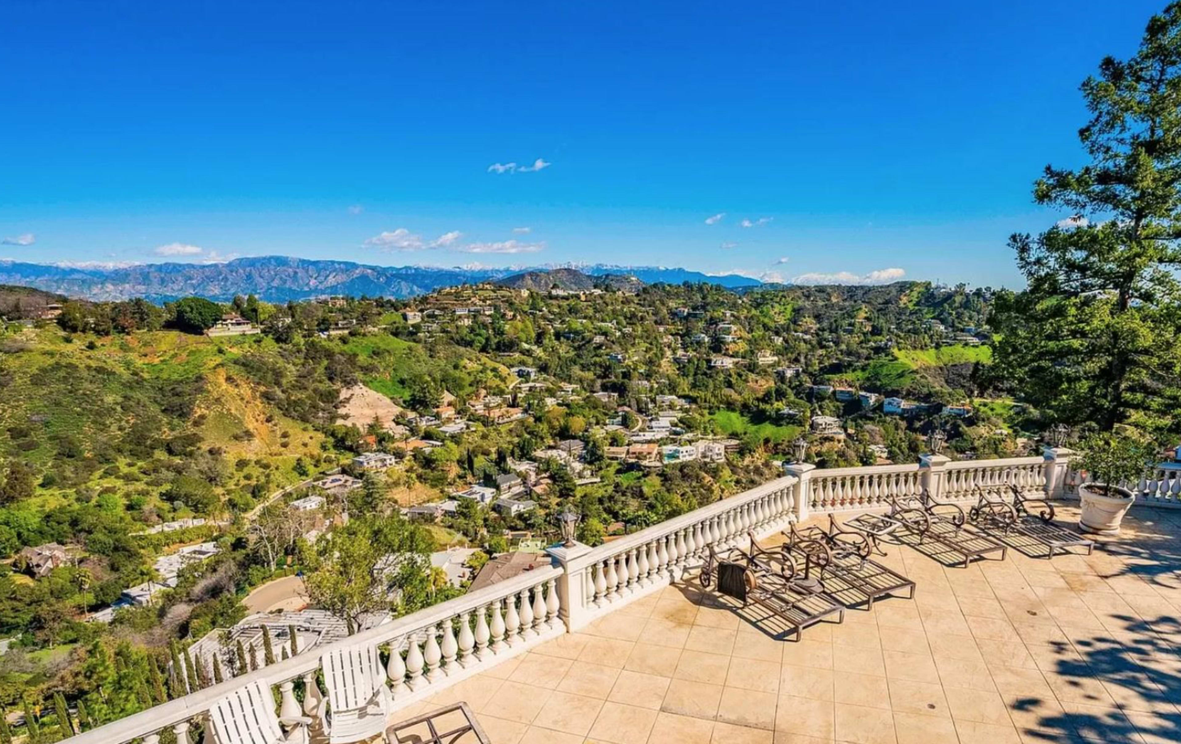 The image shows a spacious terrace with a railing, overlooking a hilly landscape dotted with houses and greenery under a clear blue sky.