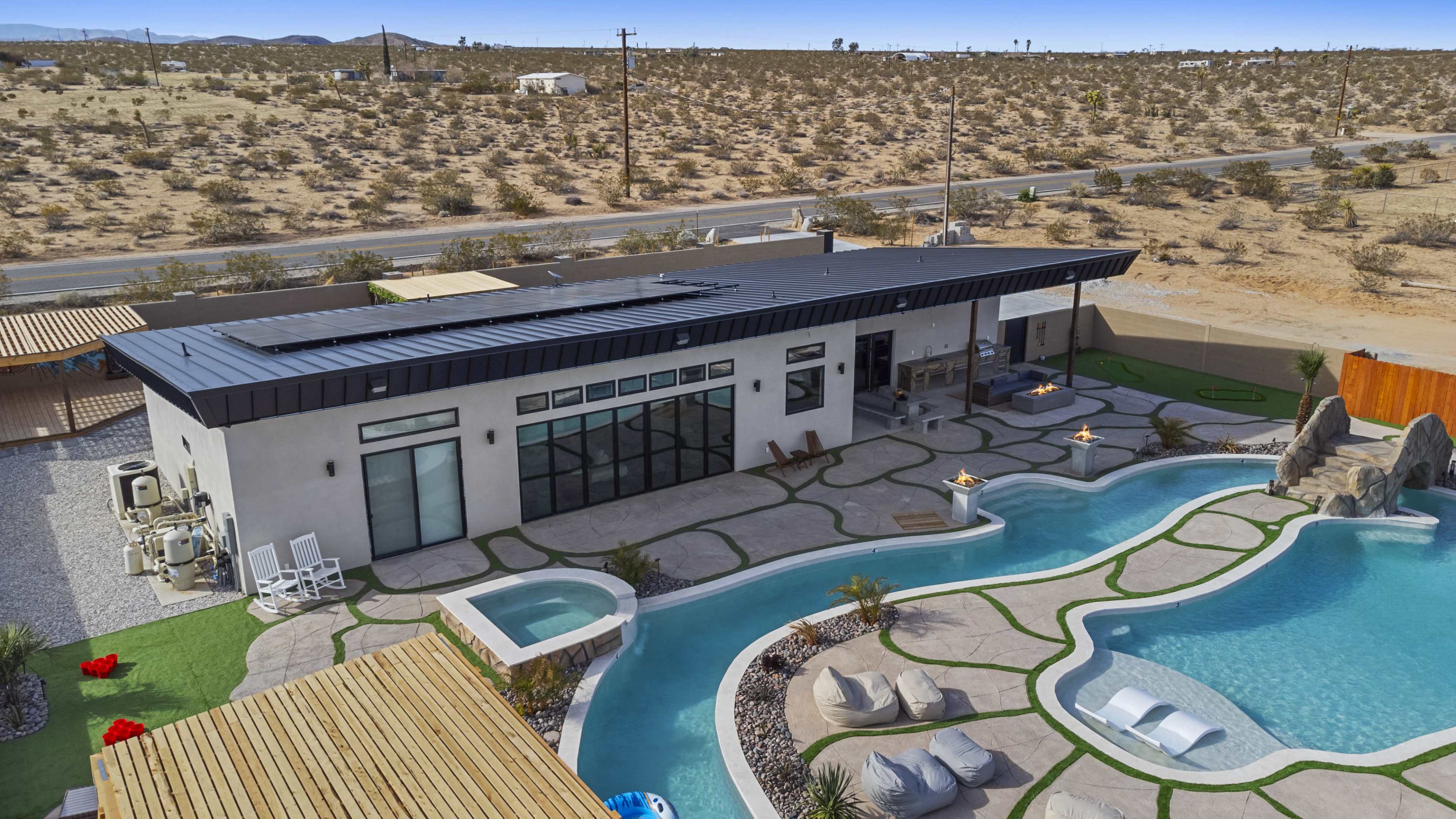 A modern house with large windows and a black roof, surrounded by a landscaped pool area and desert terrain.