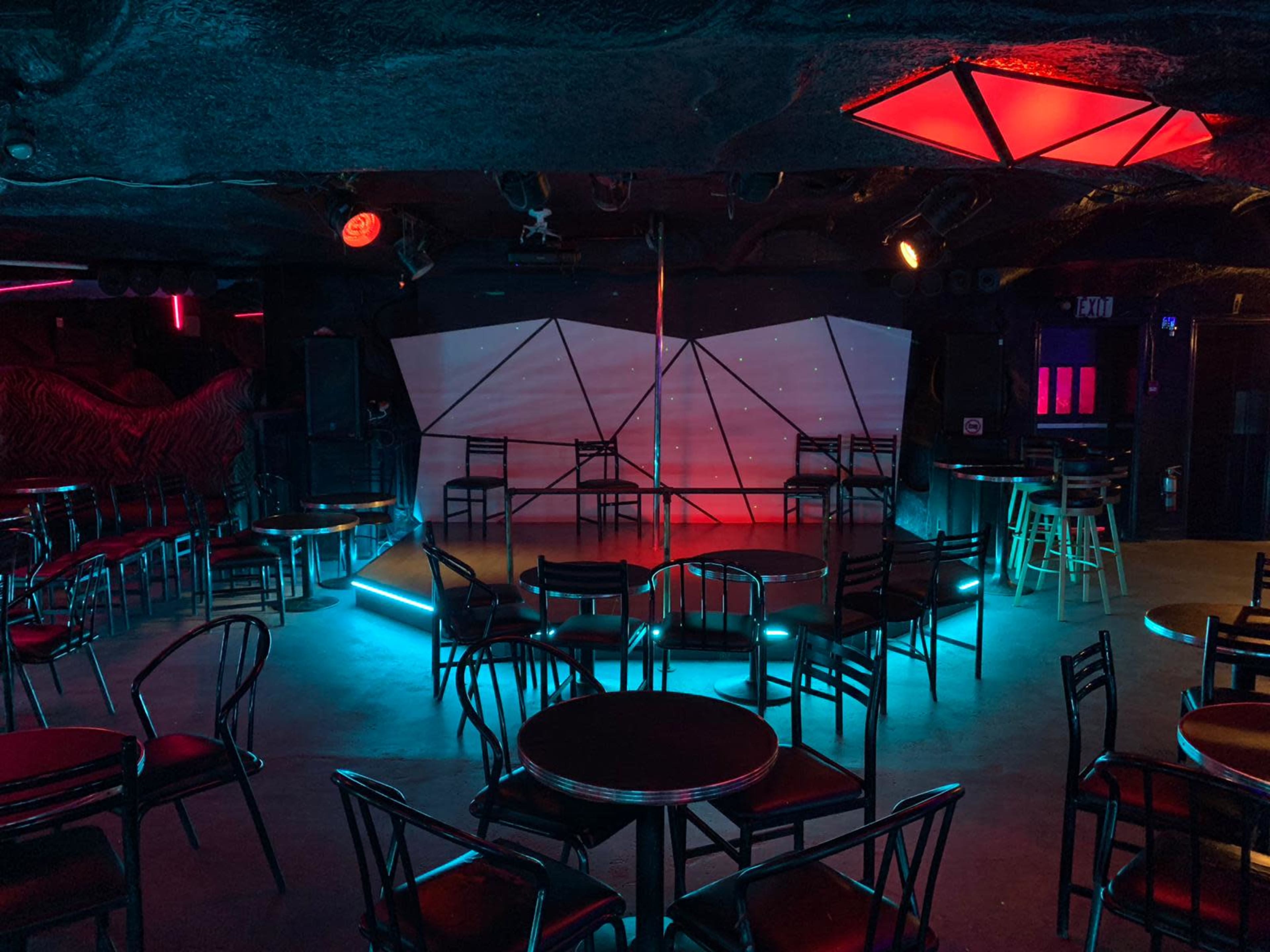 The image shows a dimly lit stage area with geometric backdrop, surrounded by empty chairs and tables in a club-like setting.