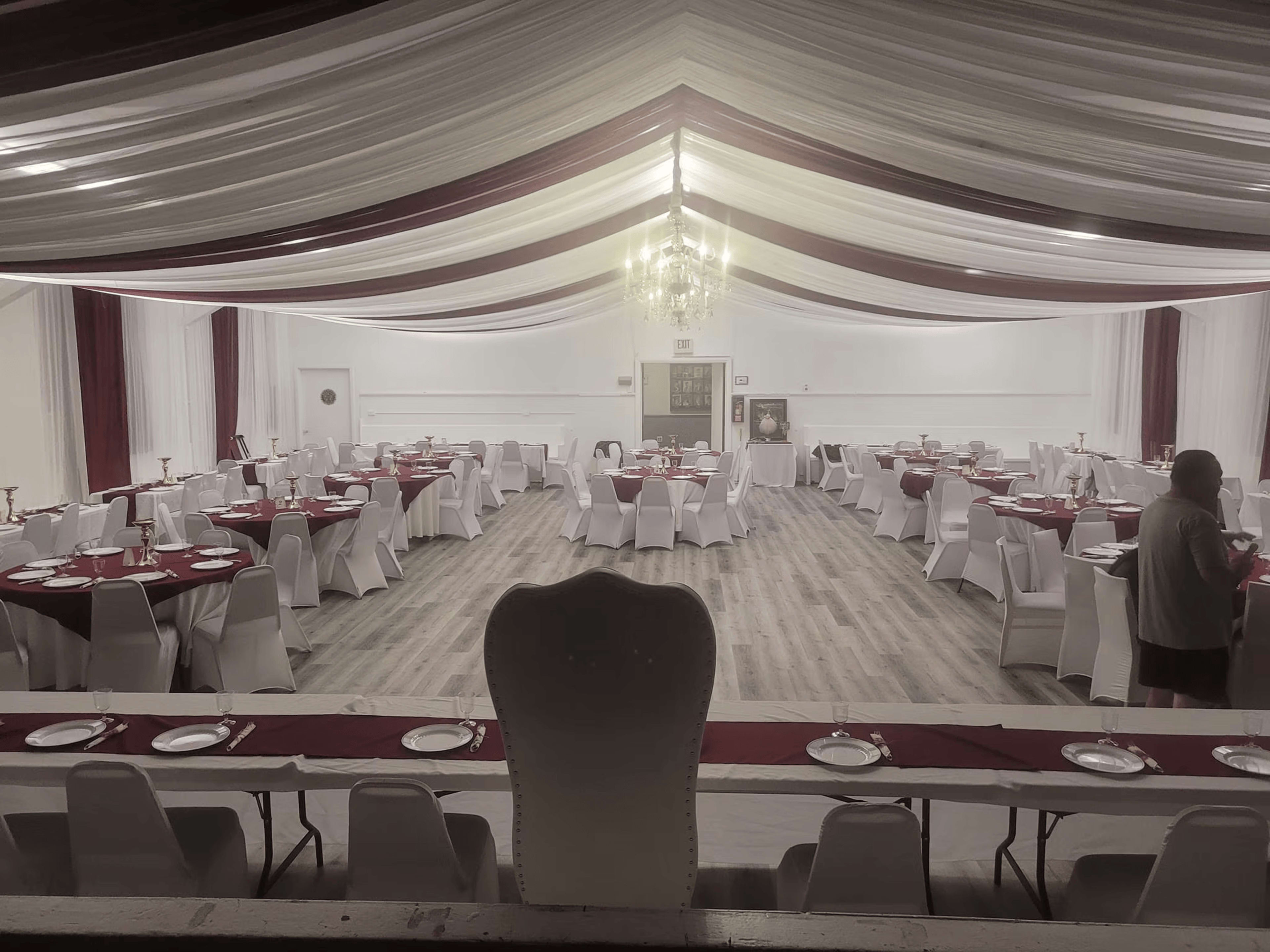A spacious banquet hall features round tables covered with red tablecloths and white chairs, with elegant drapery overhead and a chandelier in the center.