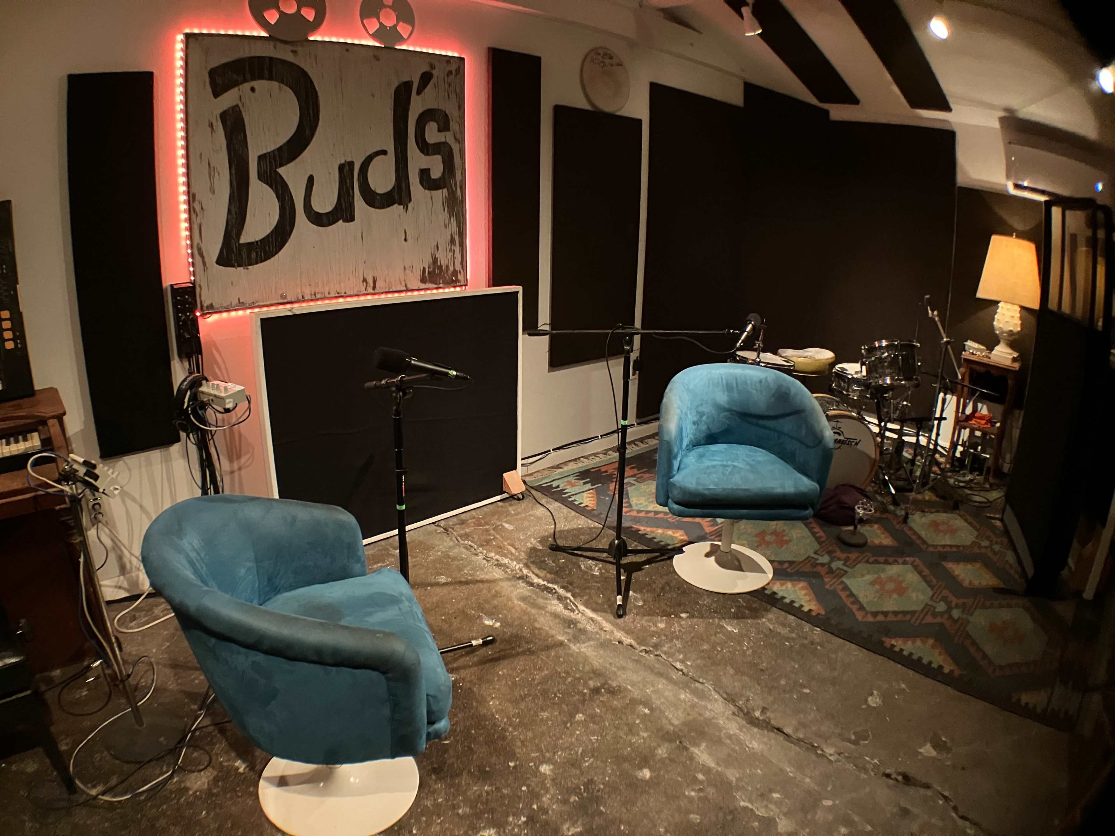 A recording studio featuring two blue chairs, a microphone setup, a drum kit, and a large "Buds" sign on the wall.