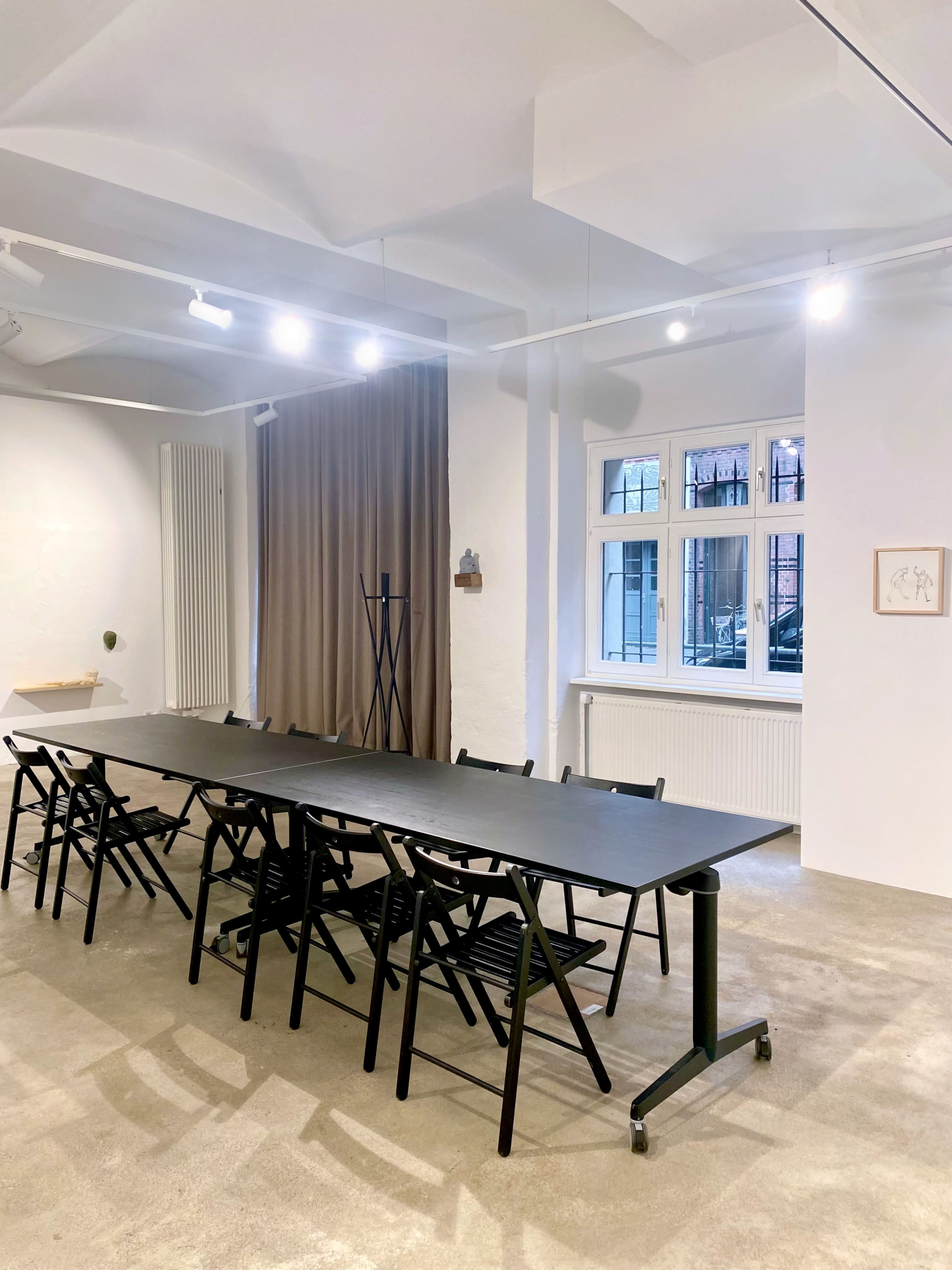 A spacious meeting room with a long black table surrounded by folding chairs, brightened by overhead lights and featuring a large window with curtains.