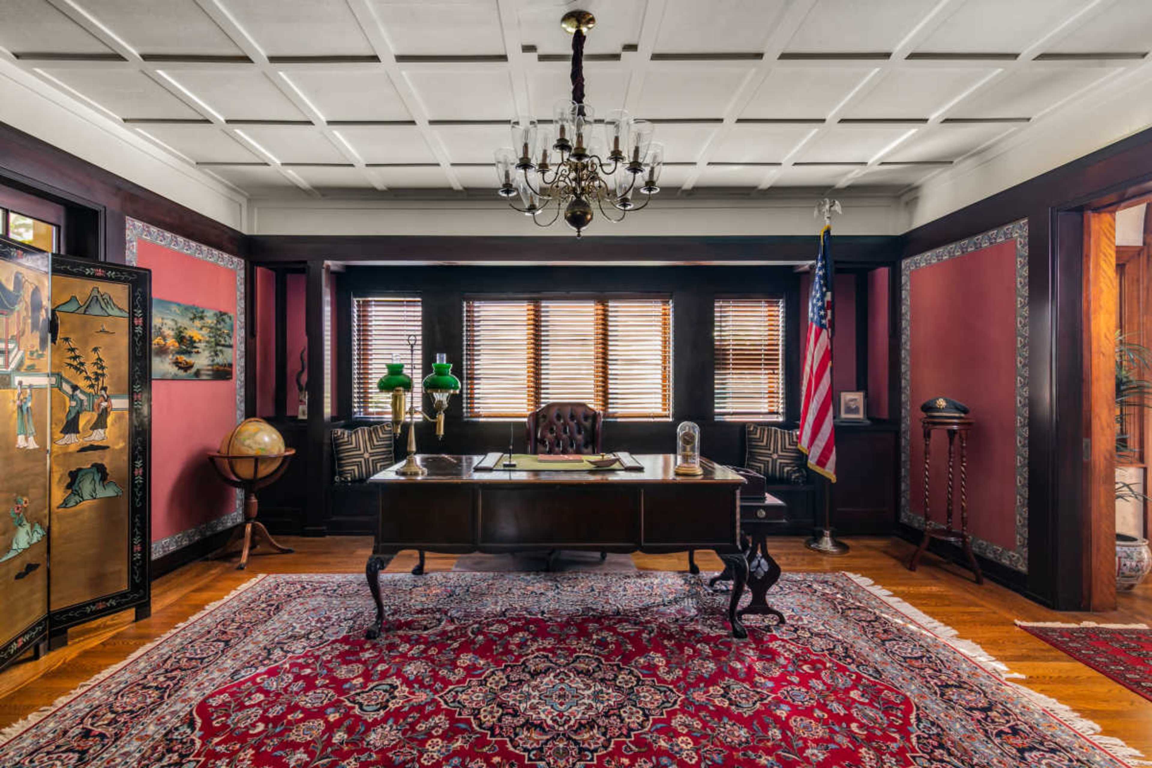 The image shows an elegantly furnished office with a large wooden desk, a globe, and various decorative elements, including an American flag and a richly patterned rug.