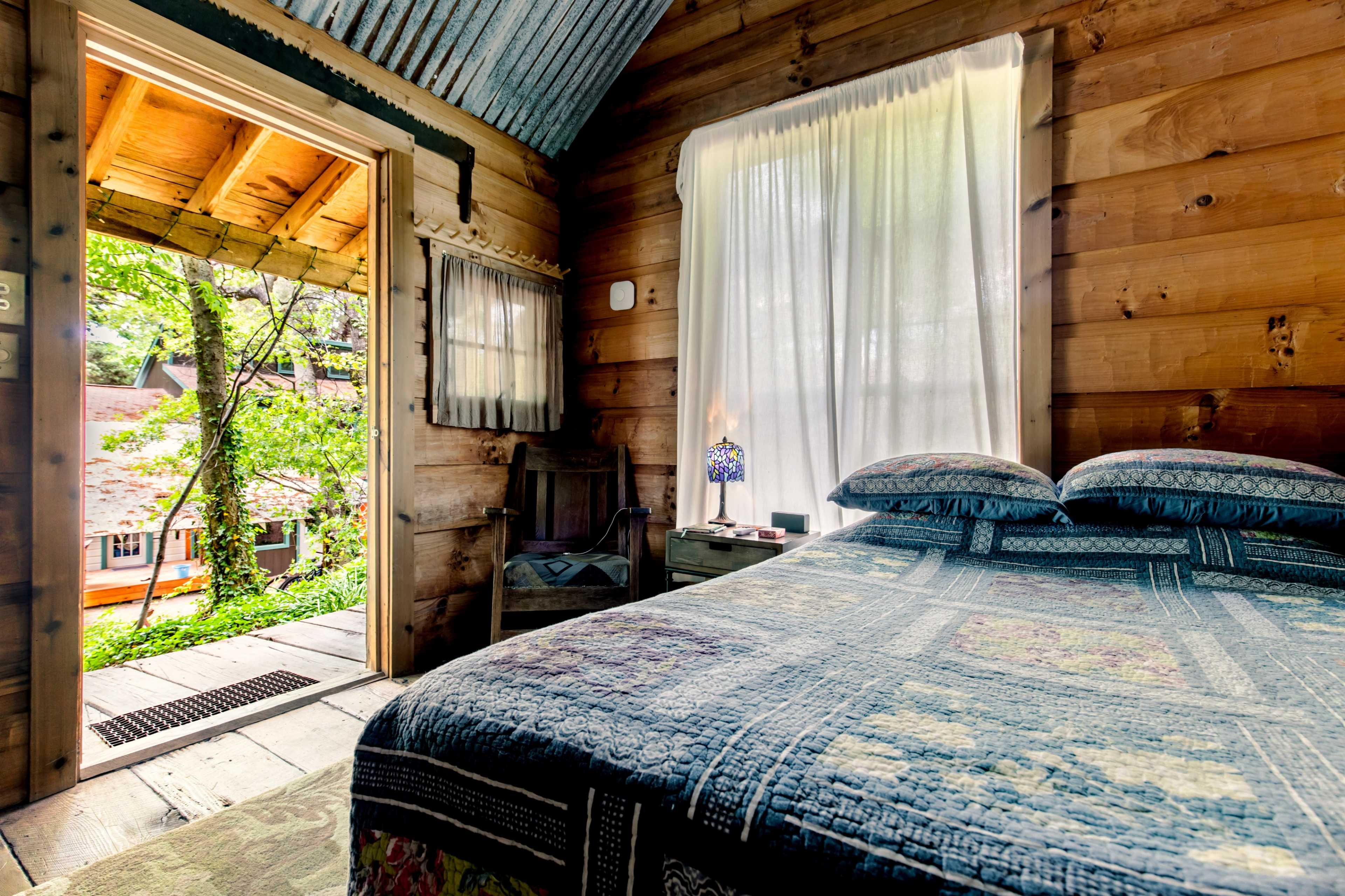 A cozy wooden bedroom features a bed with a patterned quilt, a bedside table, and an open door leading to an outdoor area surrounded by greenery.