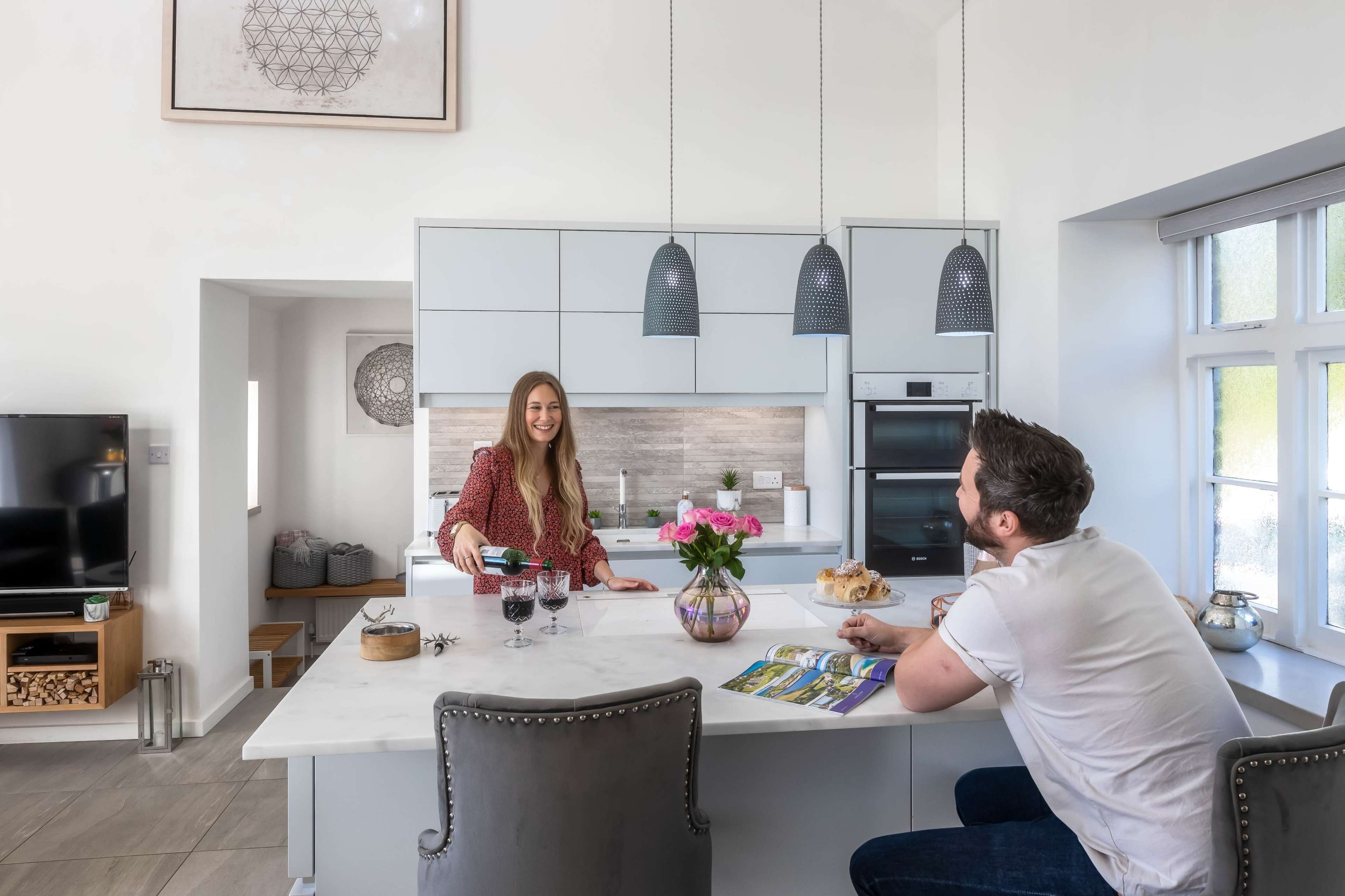 A woman serves drinks at a kitchen island while a man sits nearby, both in a modern kitchen setting.