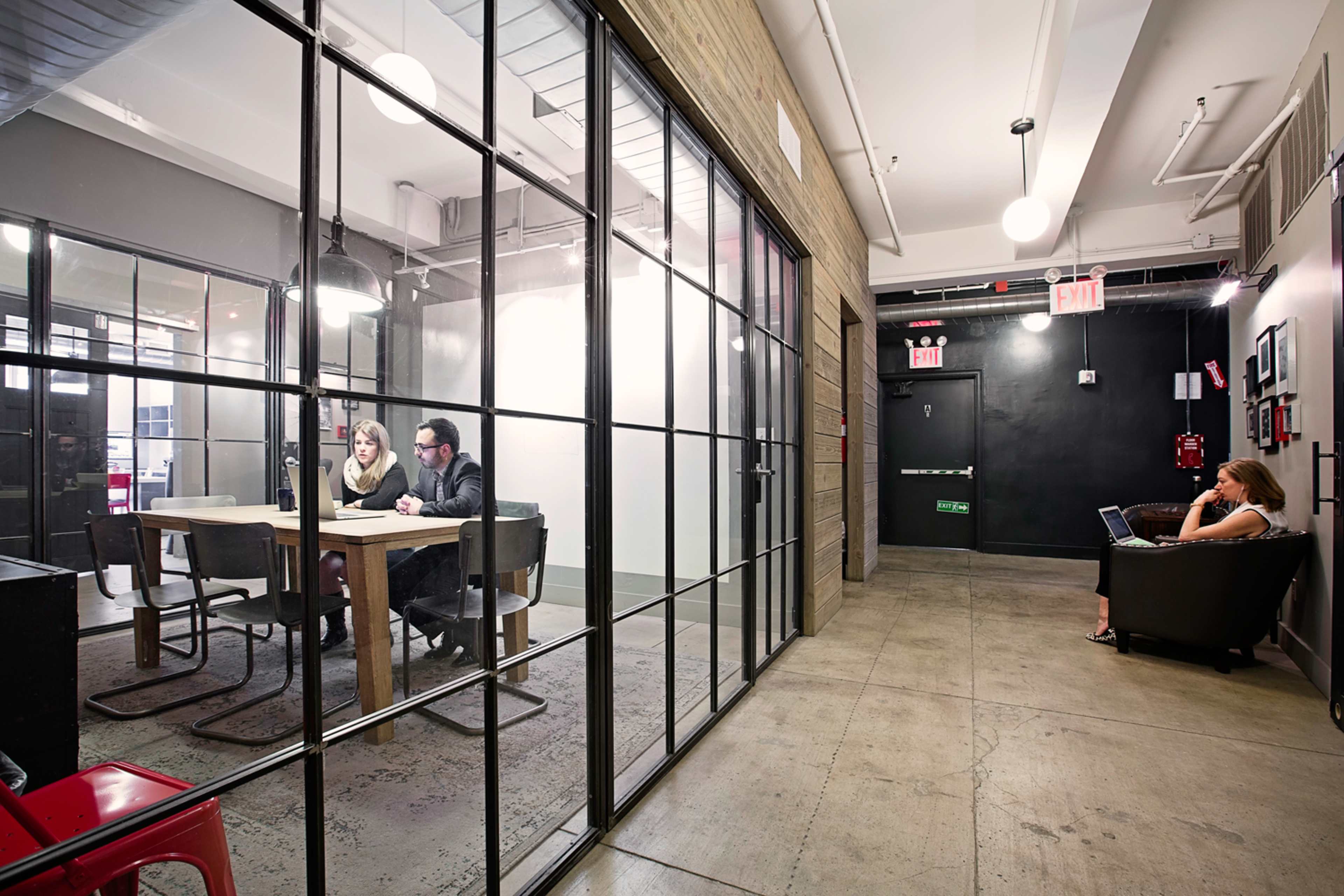 A modern office space features a glass-walled conference room with two people at a table, while a woman sits on a couch in the hallway.