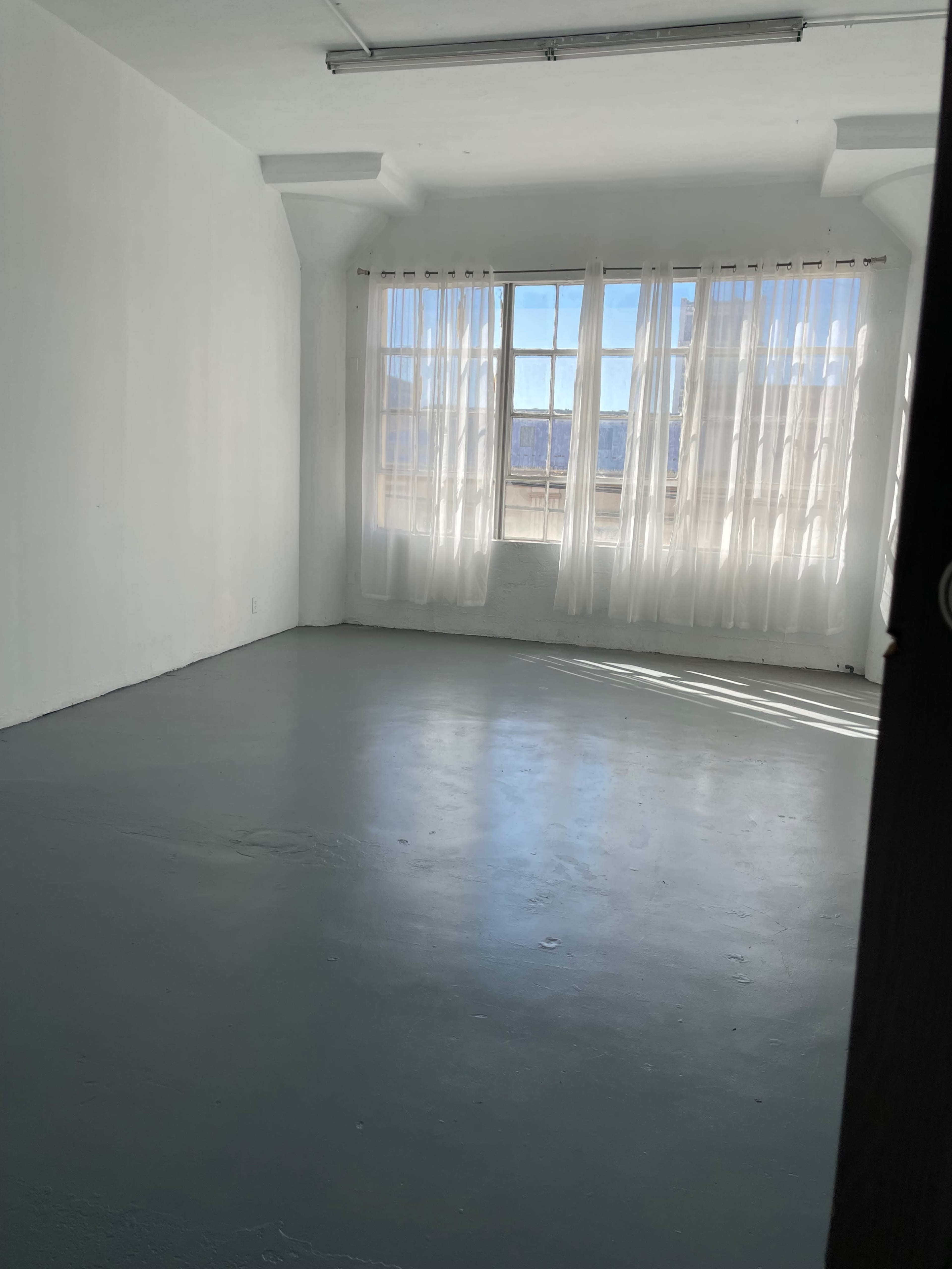 The image shows an empty room with large windows covered by sheer curtains, allowing natural light to illuminate the simple, gray floor.