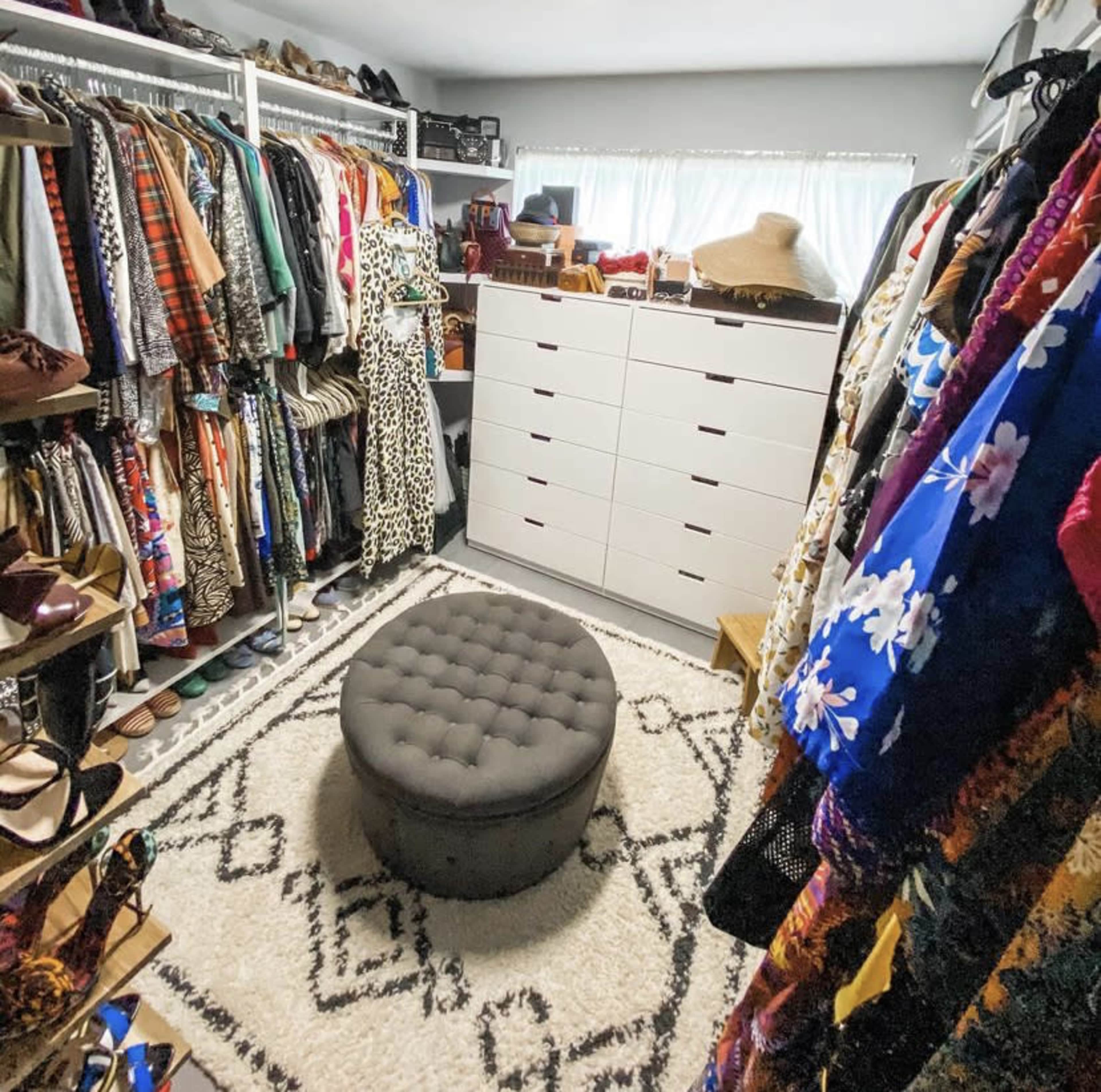A walk-in closet filled with clothes on hangers, a tufted ottoman in the center, and a white dresser against the wall.