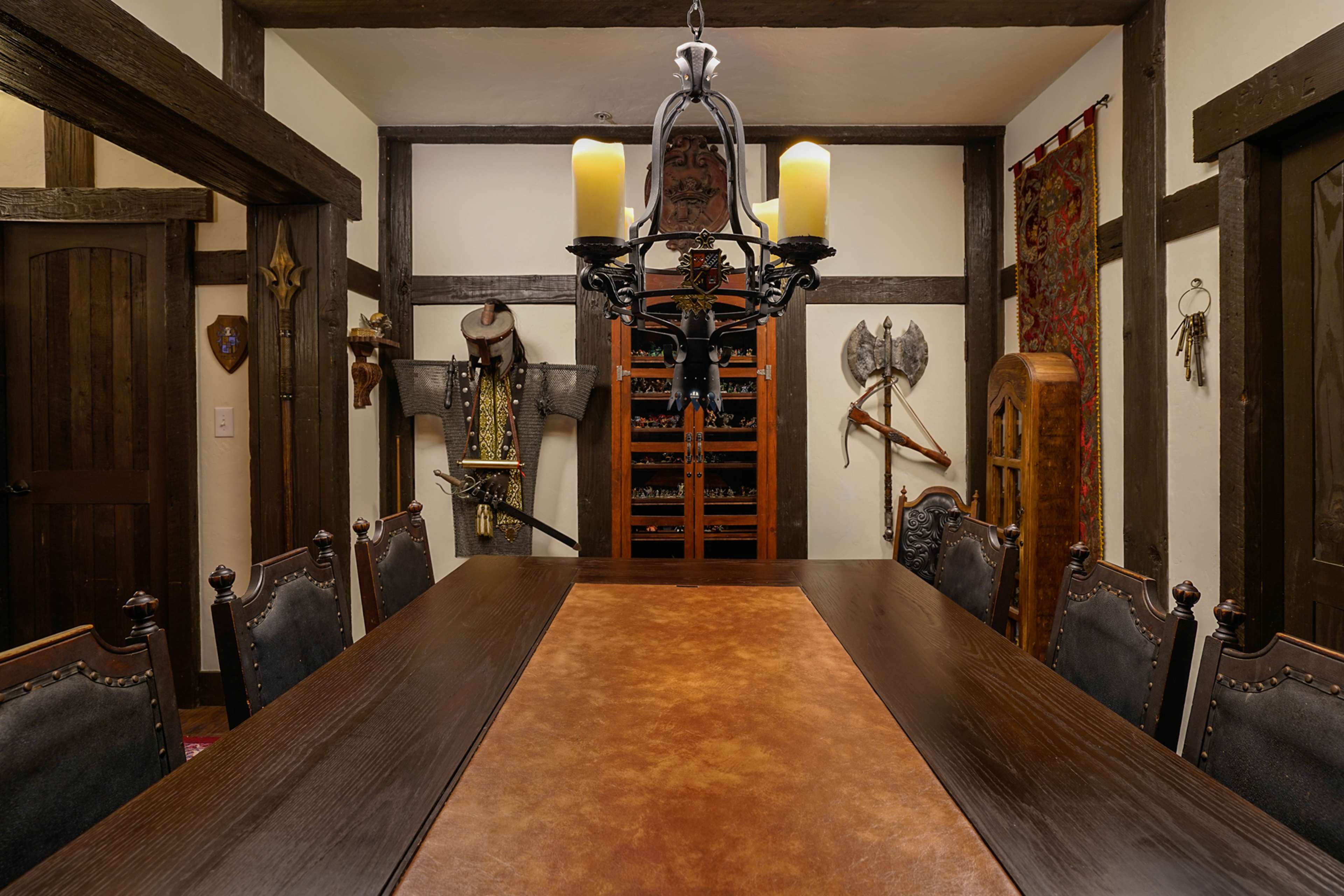 The image depicts a rustic dining room featuring a long wooden table, dark wooden beams, a wrought iron chandelier with candles, and various medieval-themed decorations on the walls.