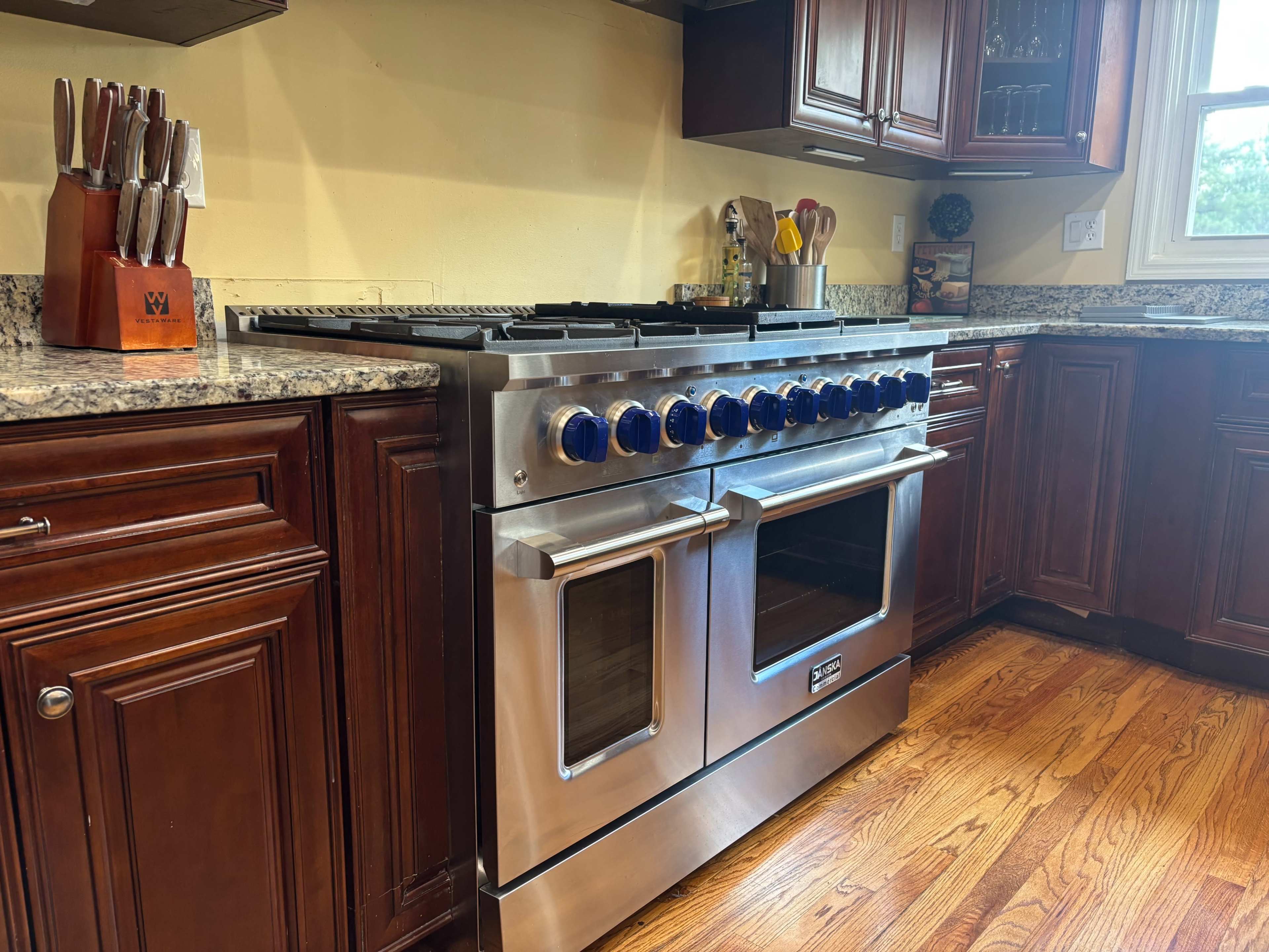 The image shows a large stainless steel gas range with blue knobs installed in a kitchen featuring dark wood cabinetry and a granite countertop.
