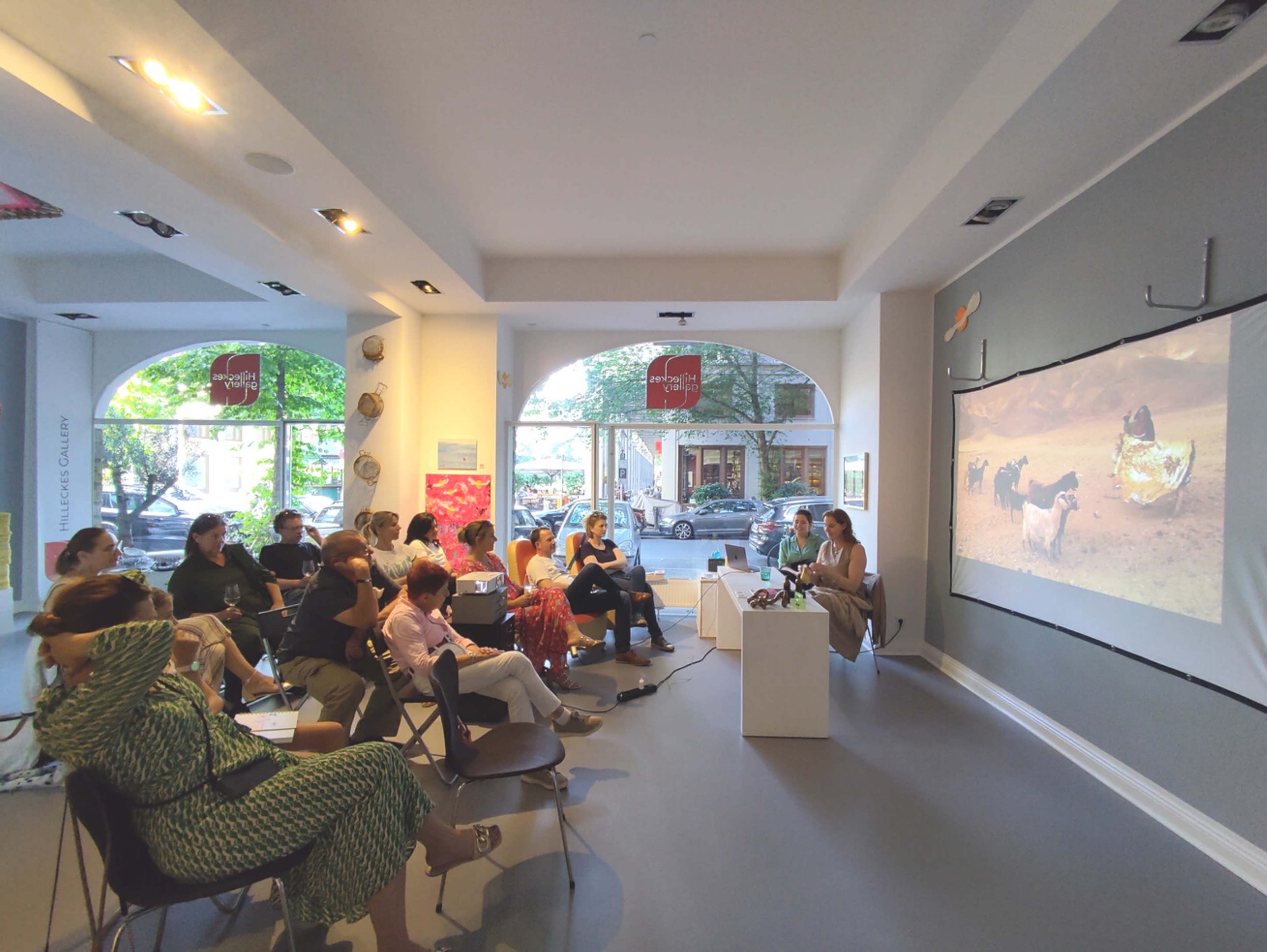 A group of people is seated in a gallery, watching a presentation that features a video projected on a wall.