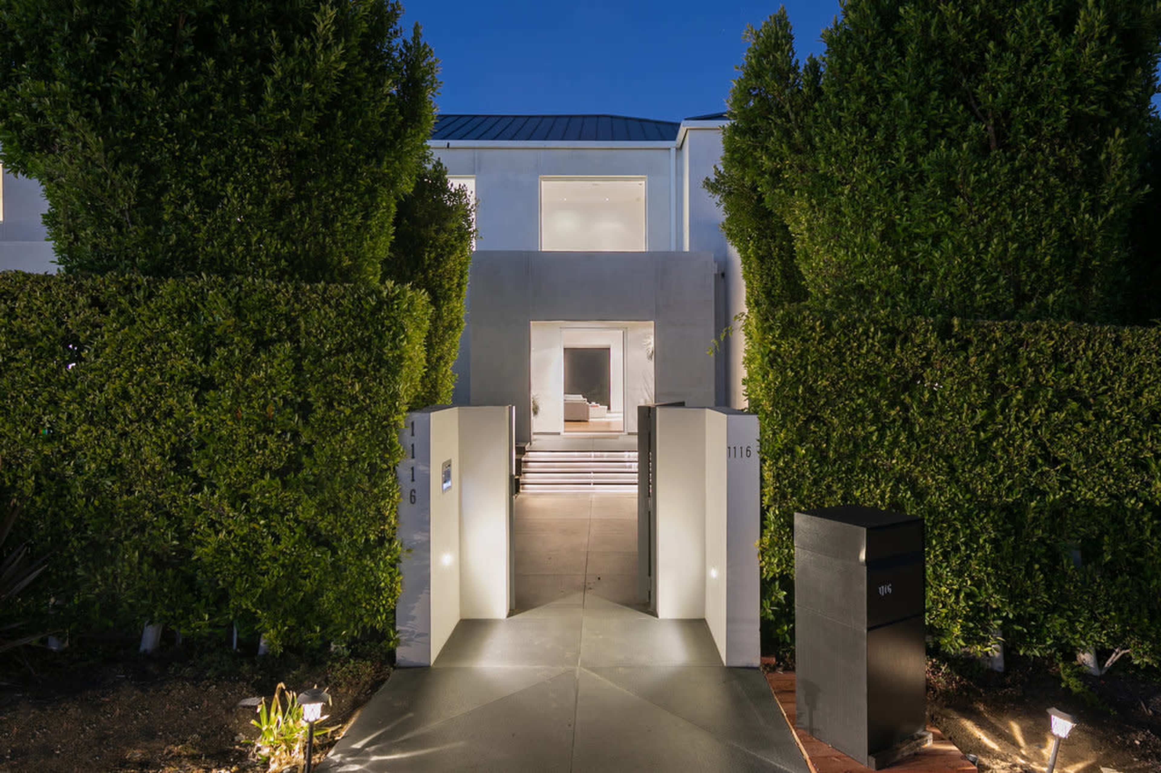 A modern house entrance framed by tall green hedges, with a pathway leading up to the illuminated front door.