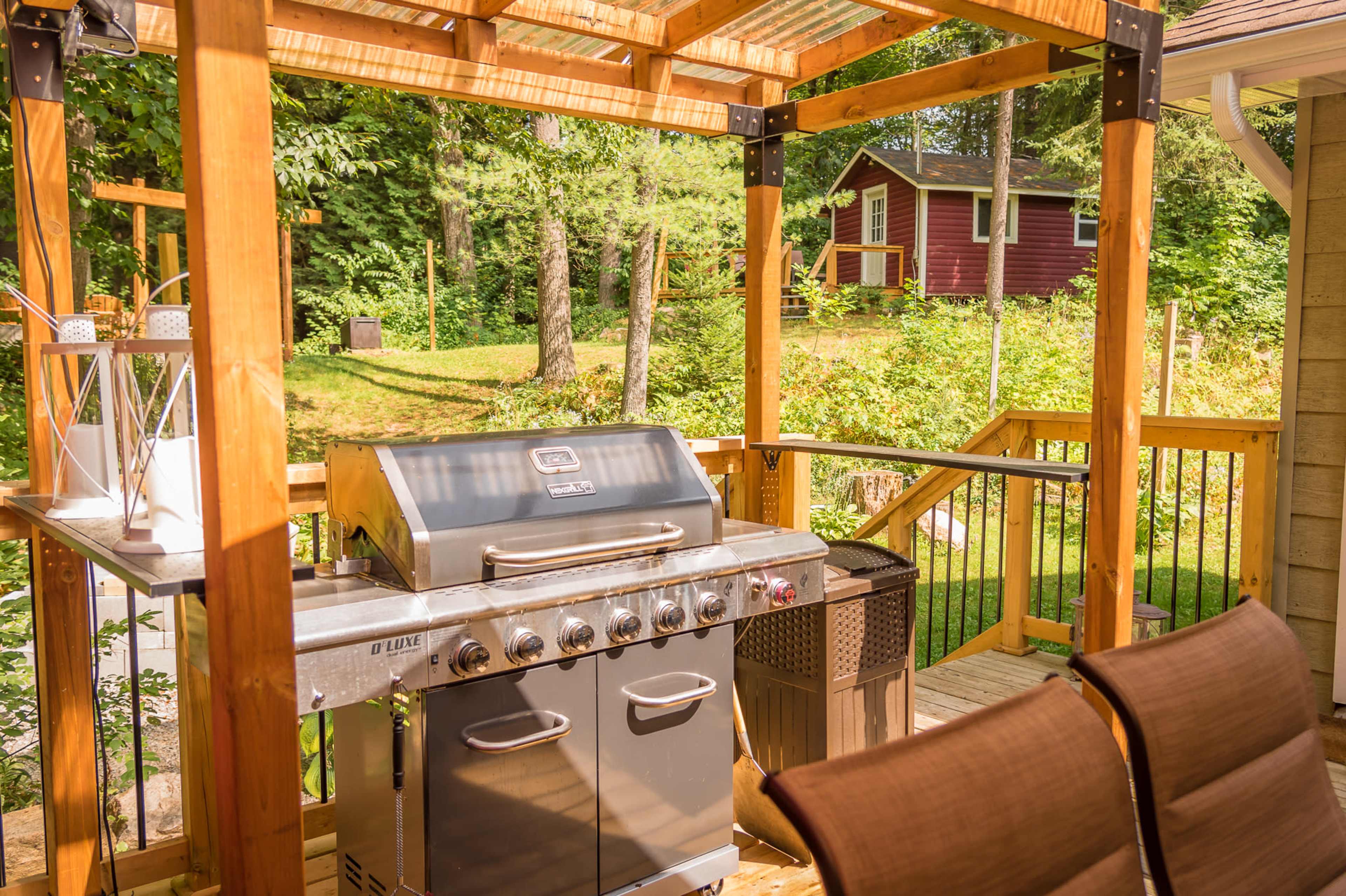 A wooden deck with a stainless steel gas grill, surrounded by greenery and a red cabin in the background.