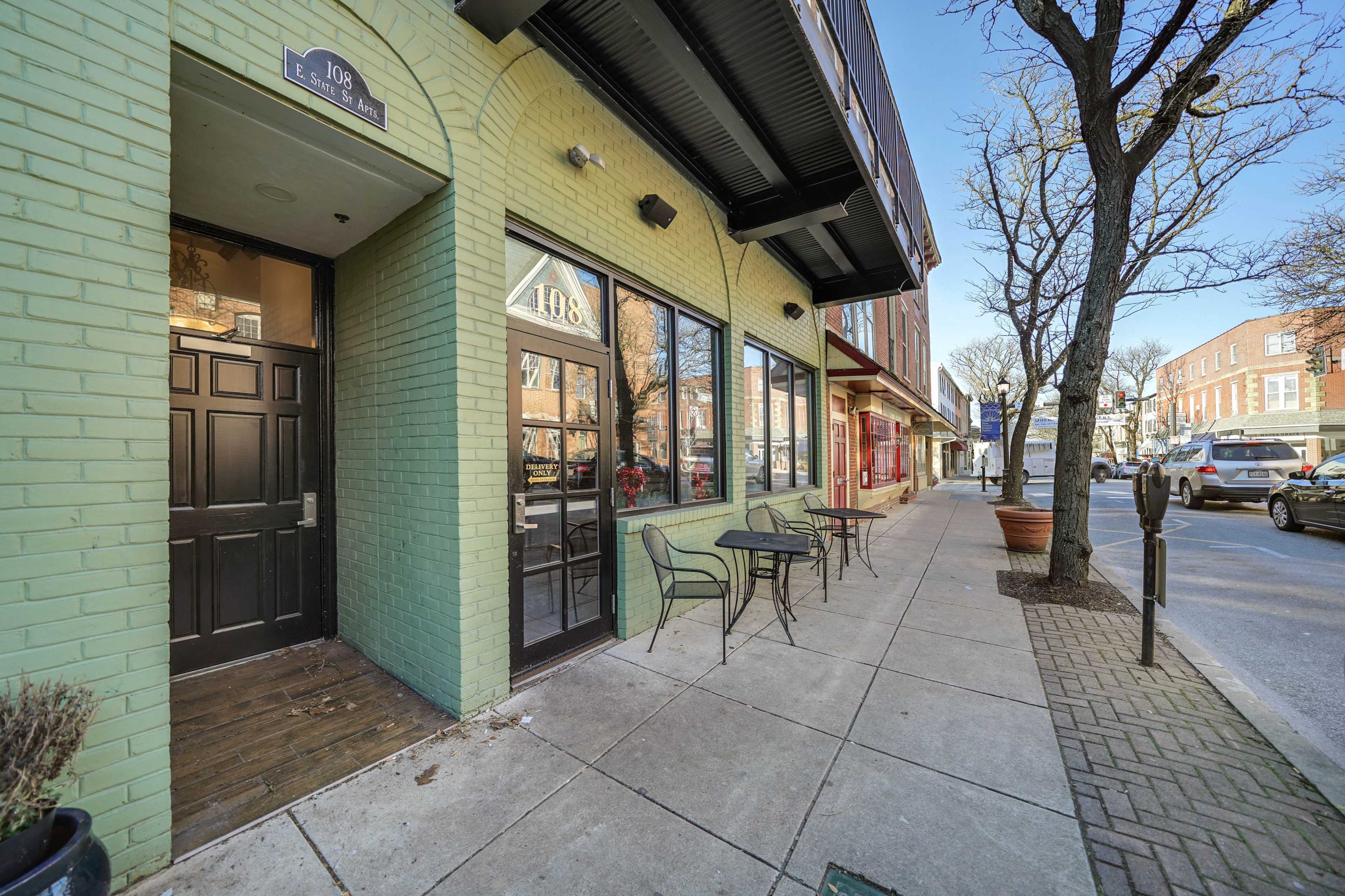 A green-brick storefront with large windows and chairs outside, located on a tree-lined sidewalk beside a street with parked cars.