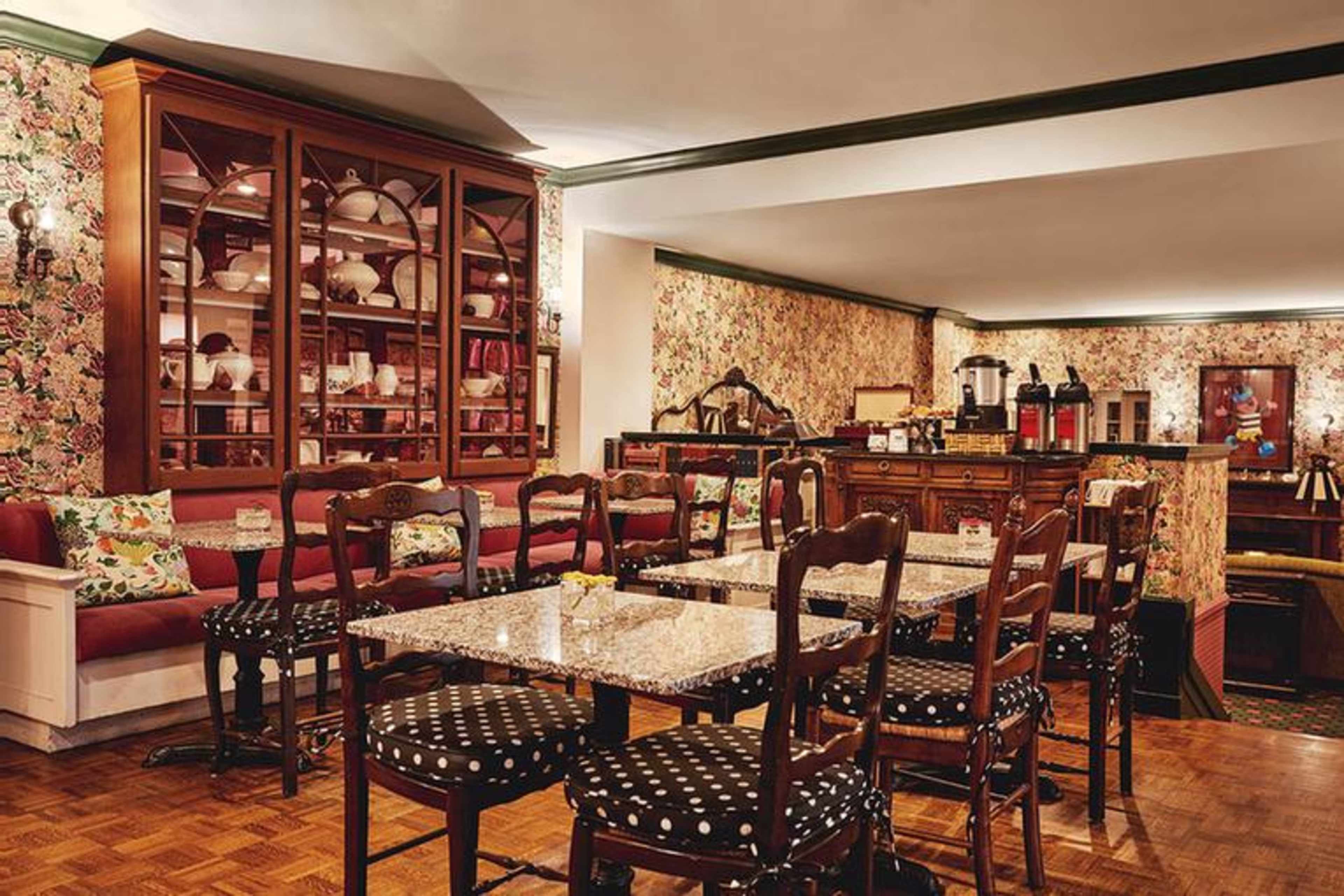 A cozy dining area with wooden tables and chairs, a floral wallpaper backdrop, and a display cabinet filled with dishware.