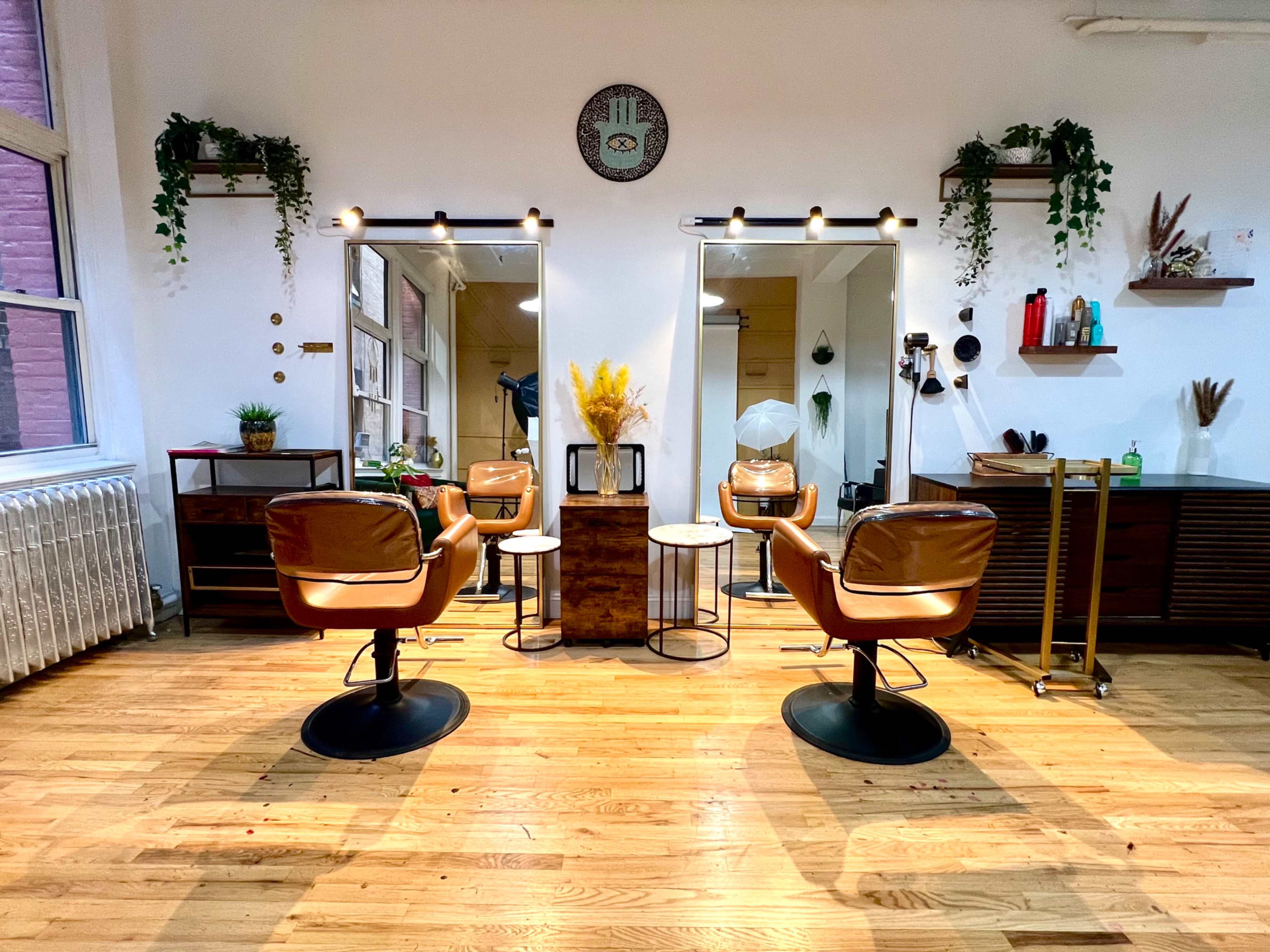 A modern hair salon with two styling stations, each equipped with a chair and mirror, set against a backdrop of decorative plants and wooden furnishings.