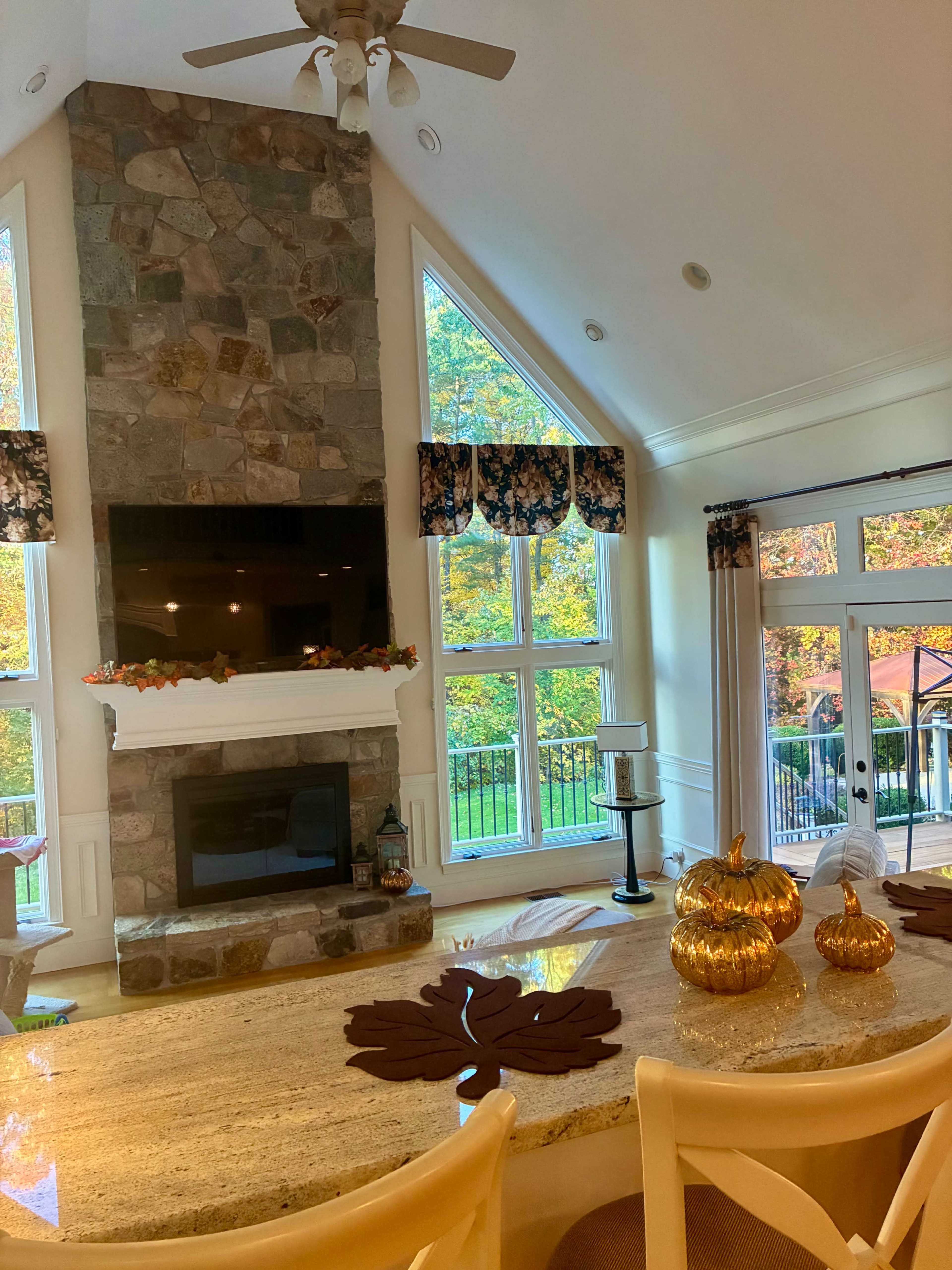The image shows a cozy living space with a stone fireplace and large windows that overlook a colorful autumn landscape, featuring decorative pumpkins on the counter.