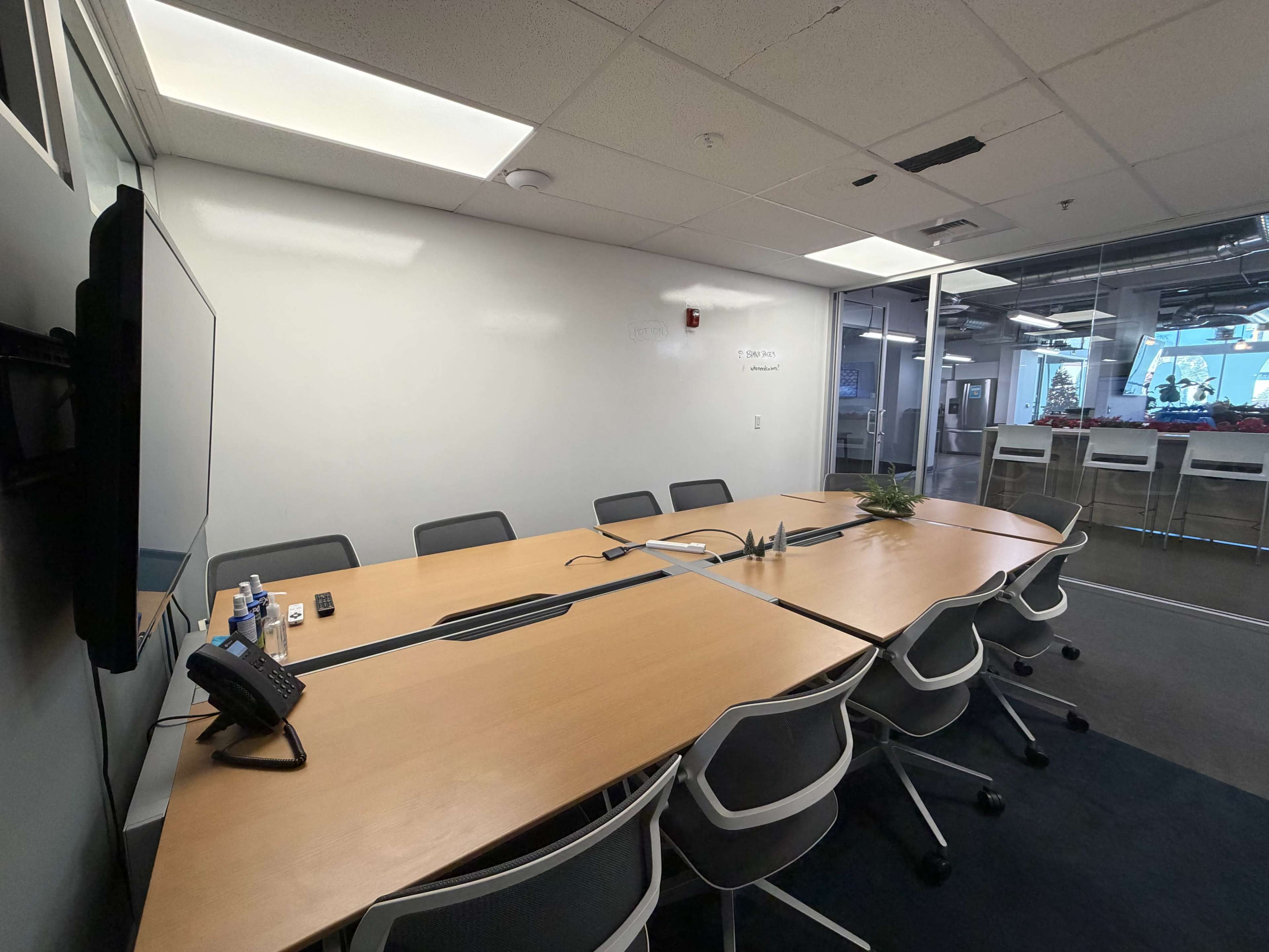 The image shows a conference room with a large wooden table surrounded by gray chairs, a television mounted on the wall, and a glass partition.