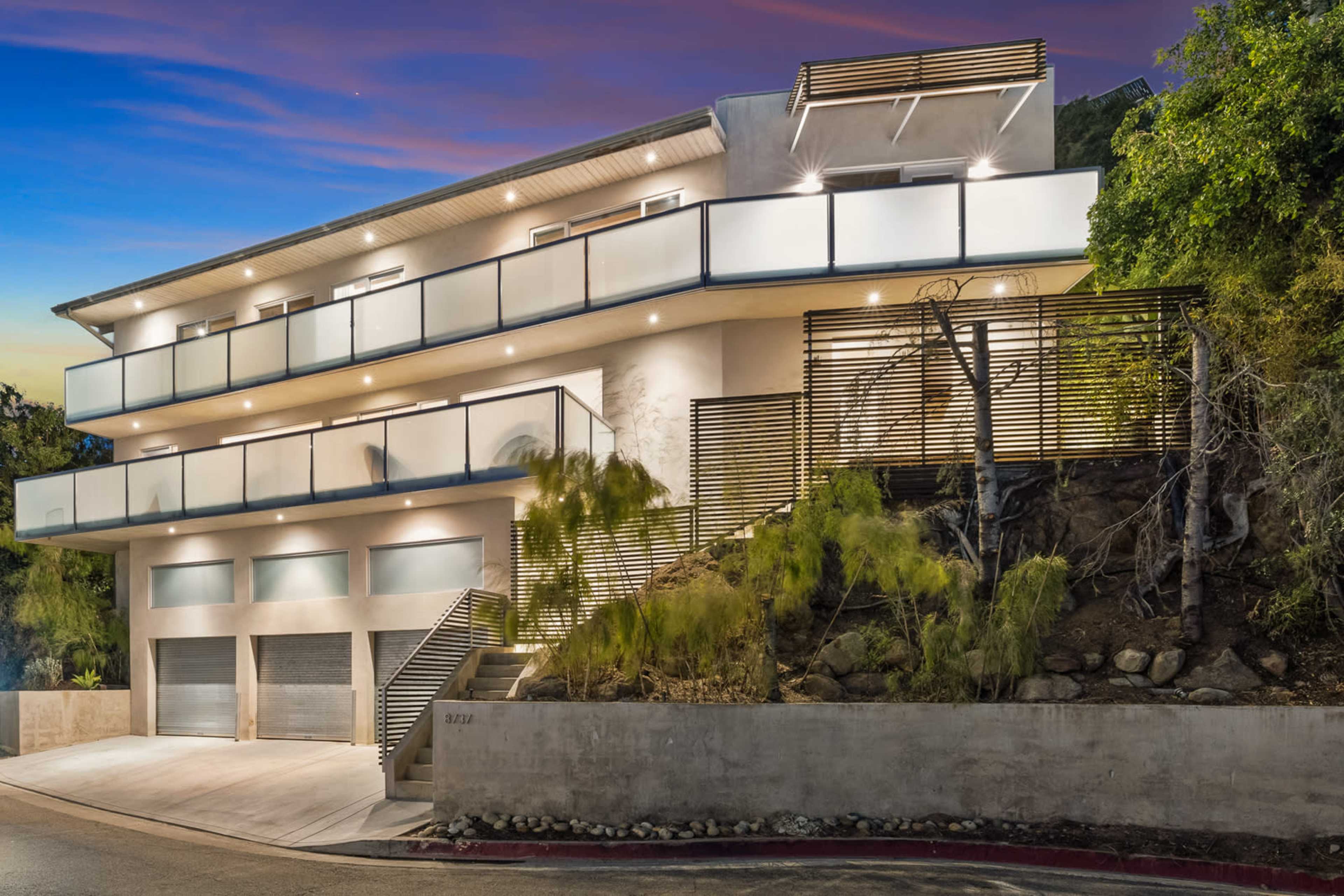 The image shows a modern multi-story house with a balcony, surrounded by foliage and featuring a paved driveway leading to two garage doors.