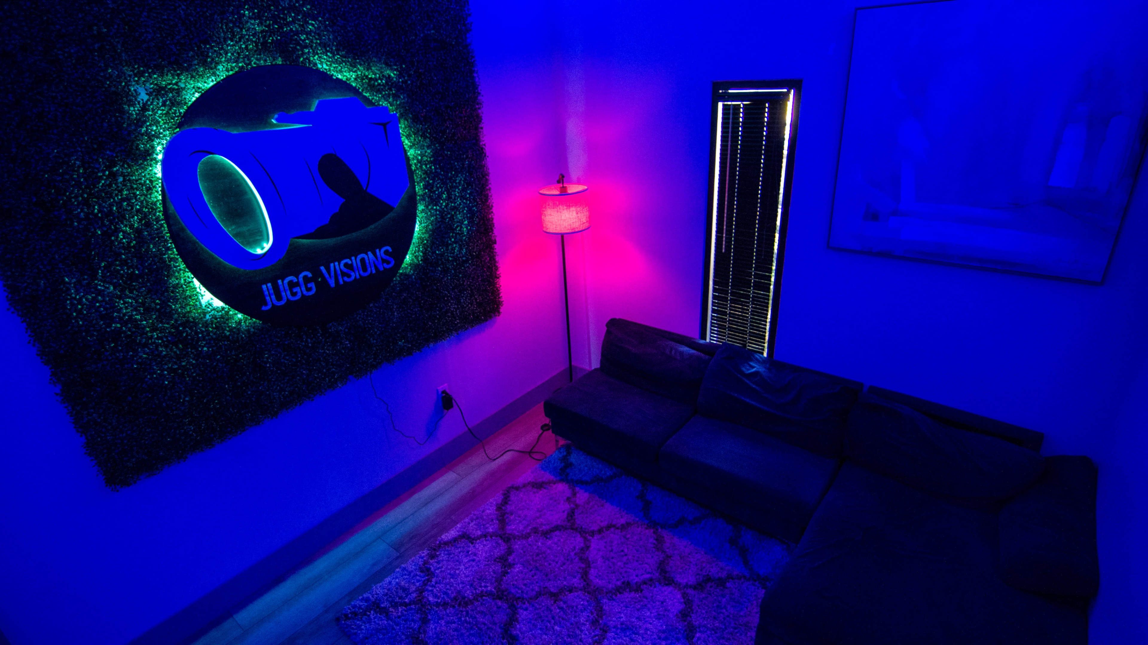 A dimly lit room with blue and pink lighting, featuring a modern couch and a decorative wall adorned with a circular sign that reads "JUGG VISIONS."
