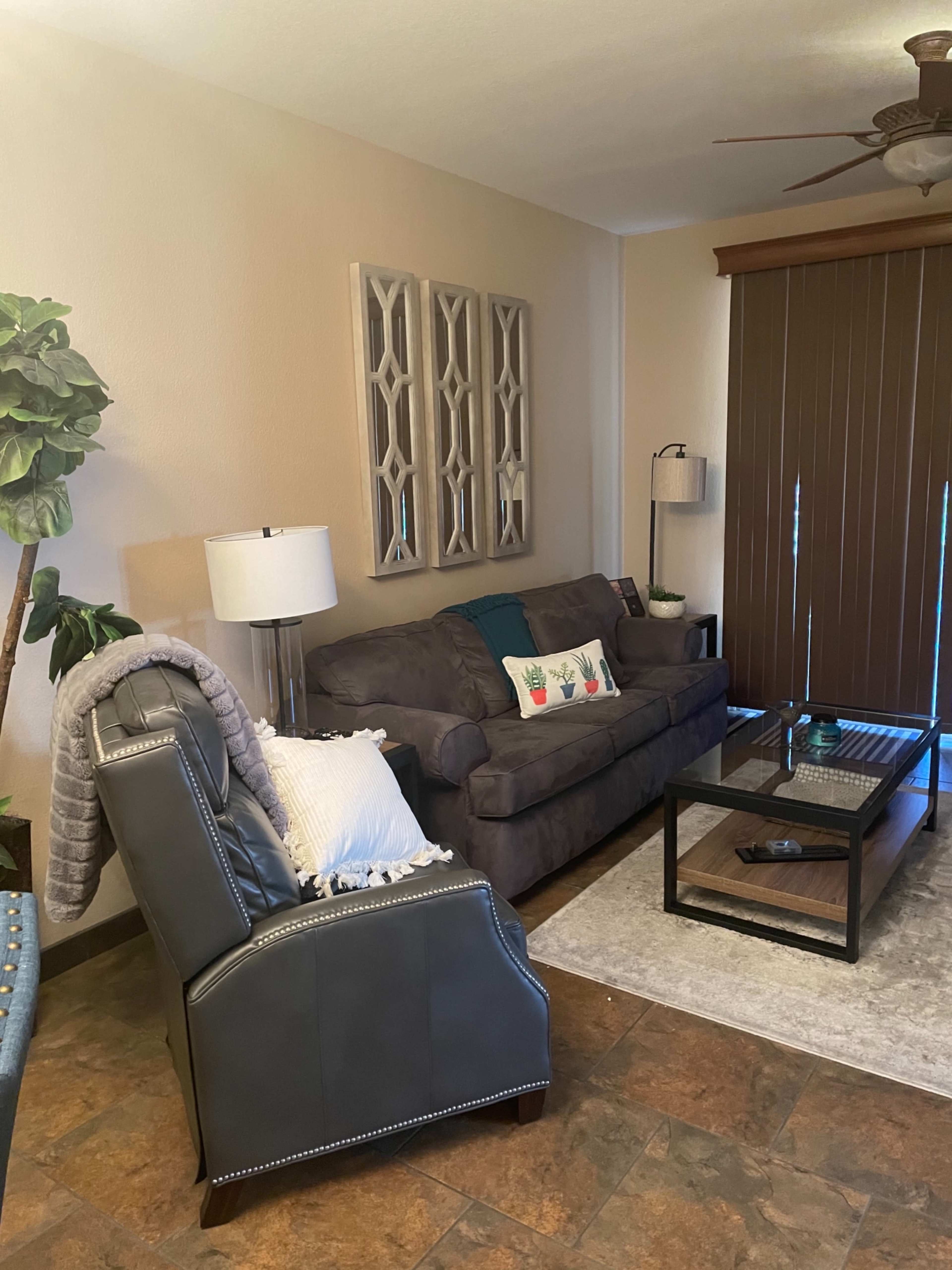 The image shows a living room with a gray sofa, a matching chair, a glass coffee table, and decorative wall art.
