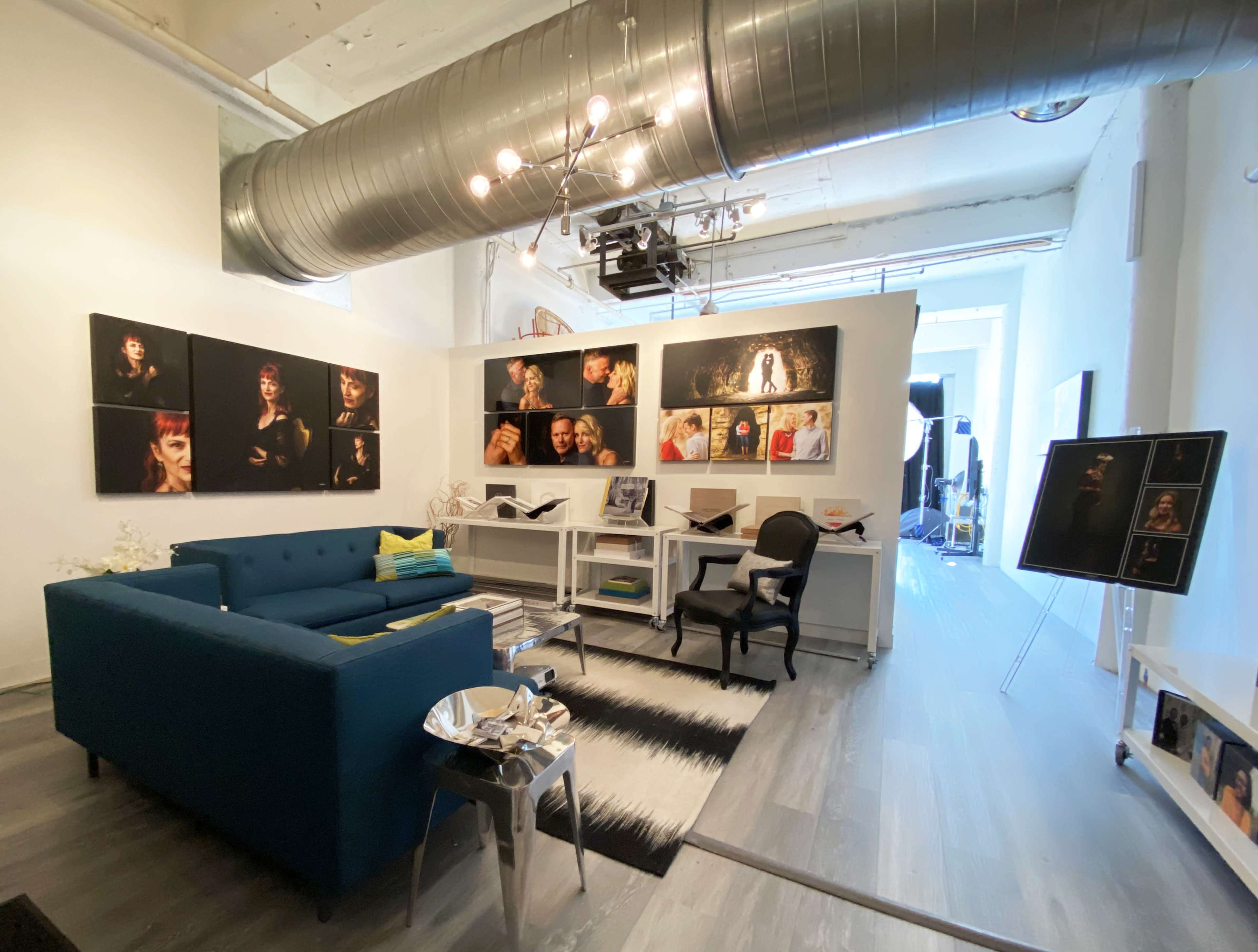 The image shows a modern studio space featuring a blue couch, white desks with chairs, and several framed photographs displayed on the walls.