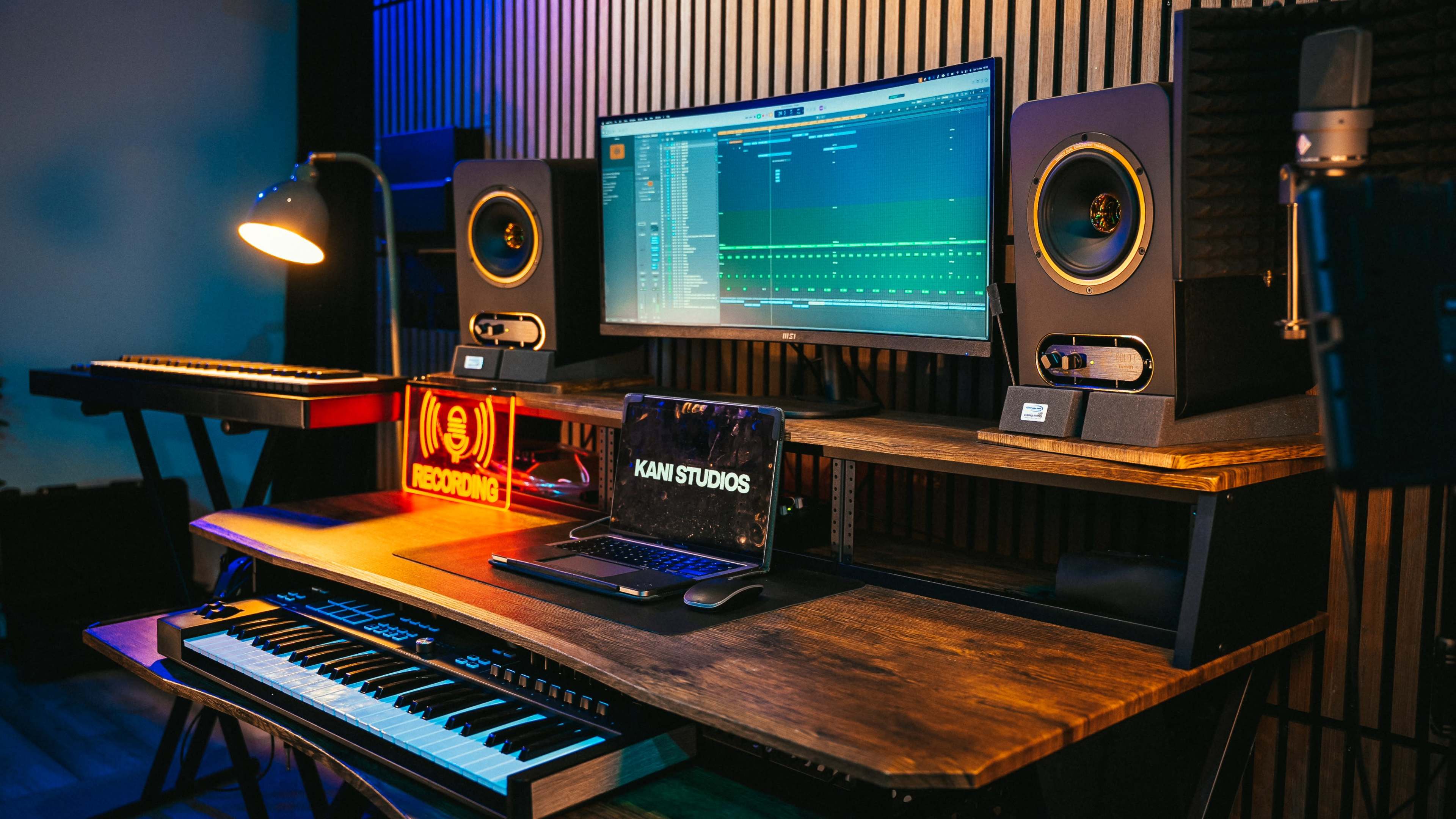 The image shows a modern music production studio featuring a computer setup with audio software displayed on a large monitor, flanked by studio monitors and a keyboard, all arranged on a wooden desk.