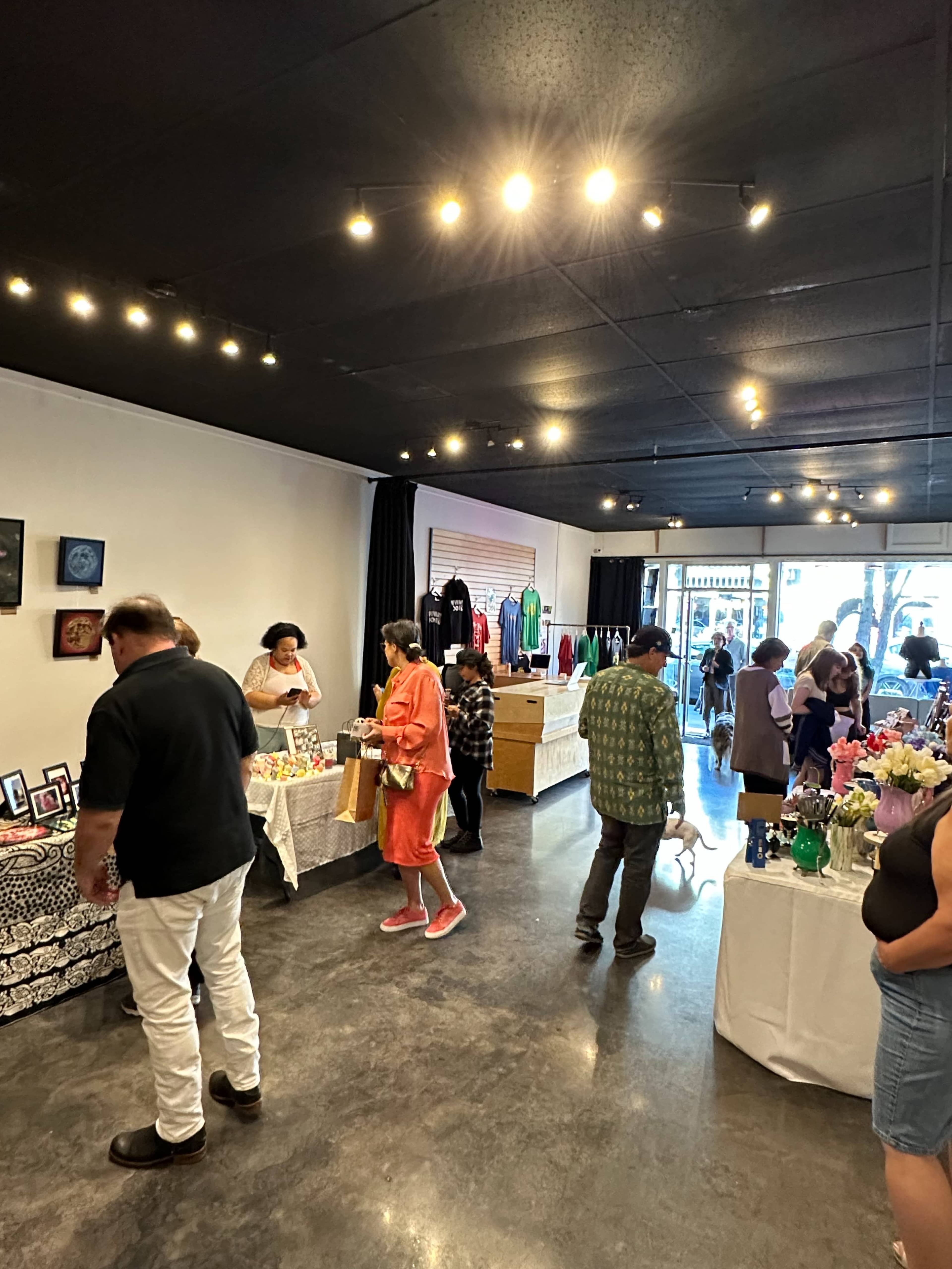 A busy boutique space with several people browsing merchandise displayed on tables, including clothing and artwork.