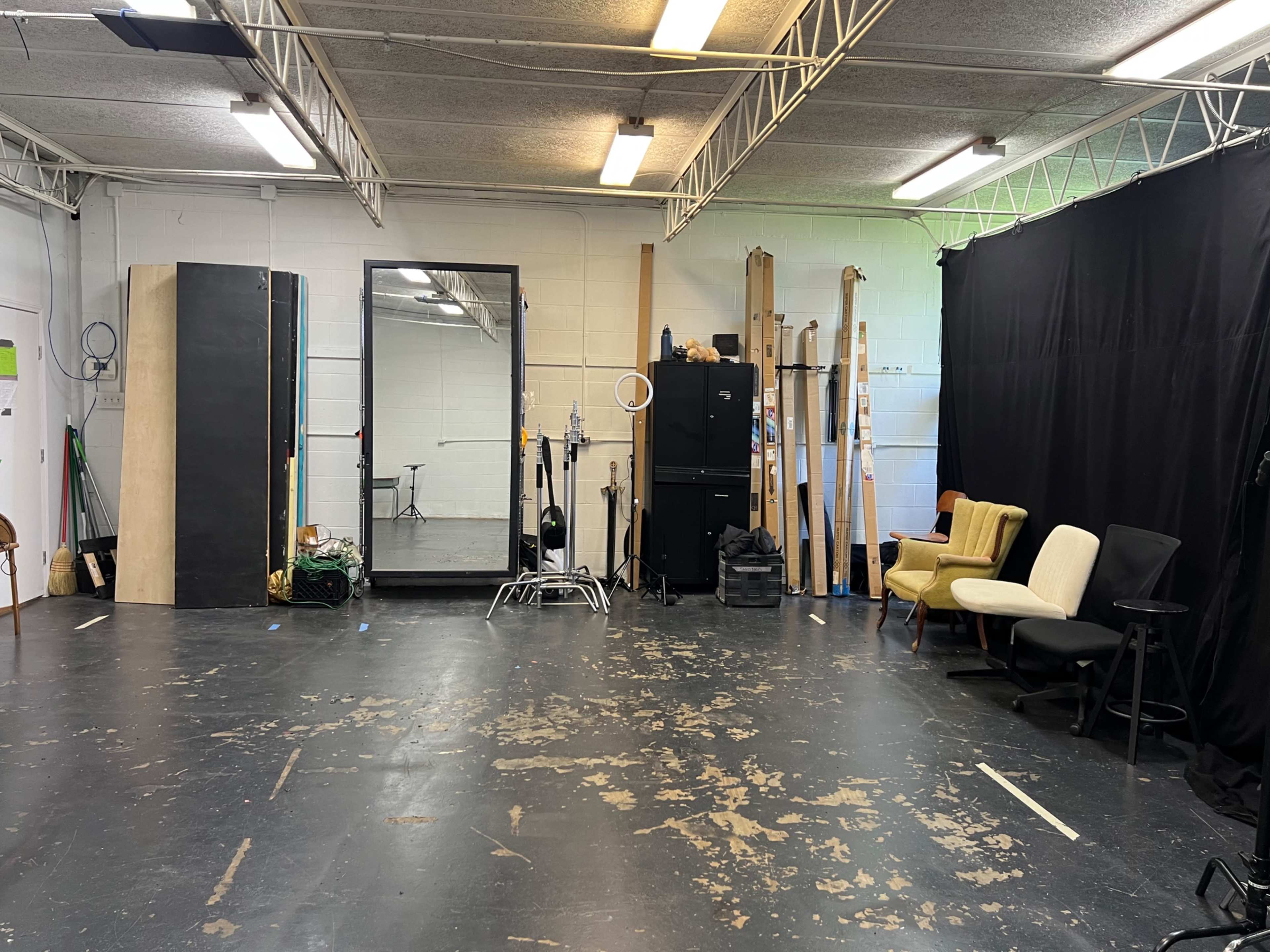 The image shows an empty studio space featuring a large mirror, various chairs, and wooden stands against the walls.