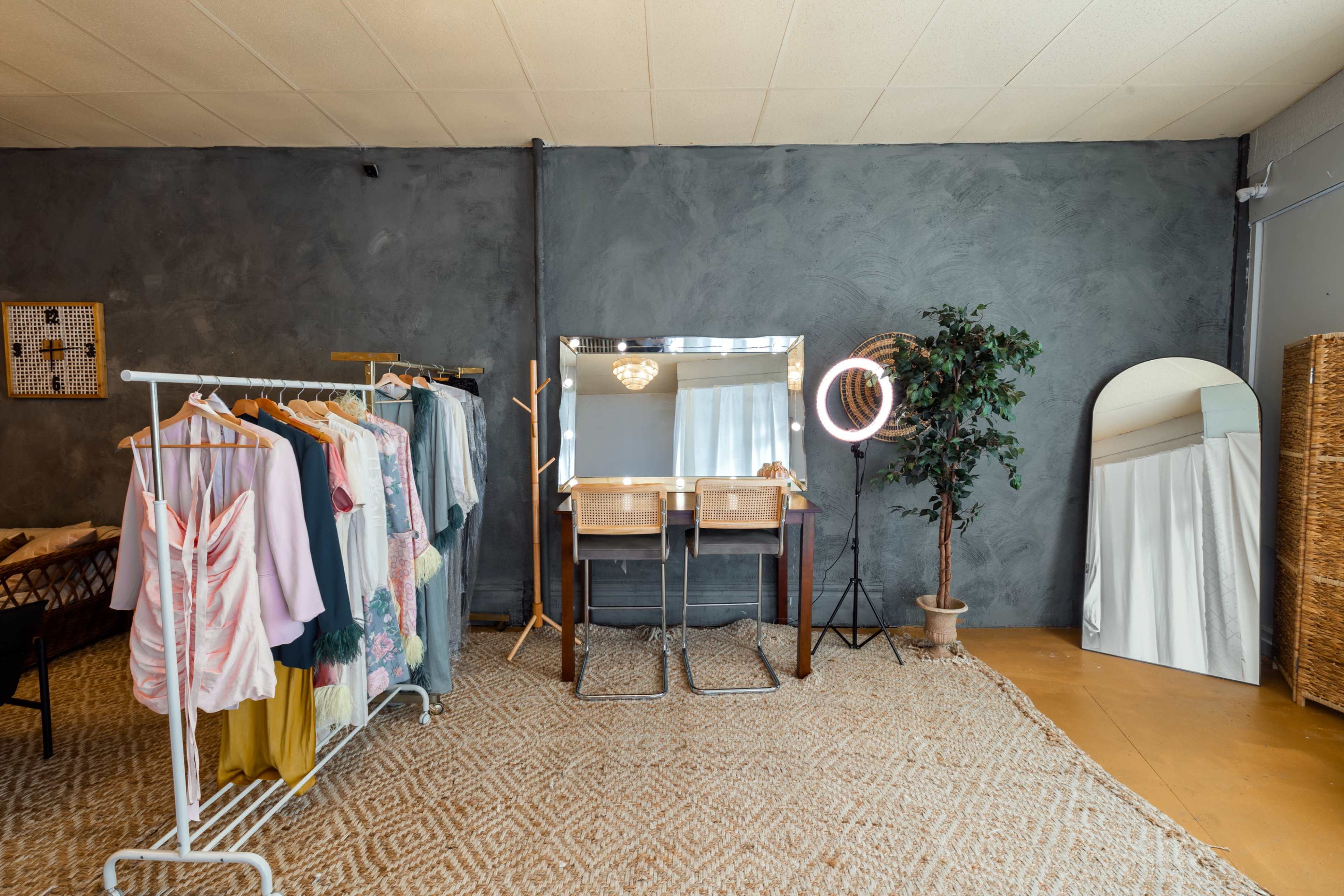 A boutique interior featuring a clothing rack, a vanity mirror with lights, a potted plant, and a large mirror against a gray wall.