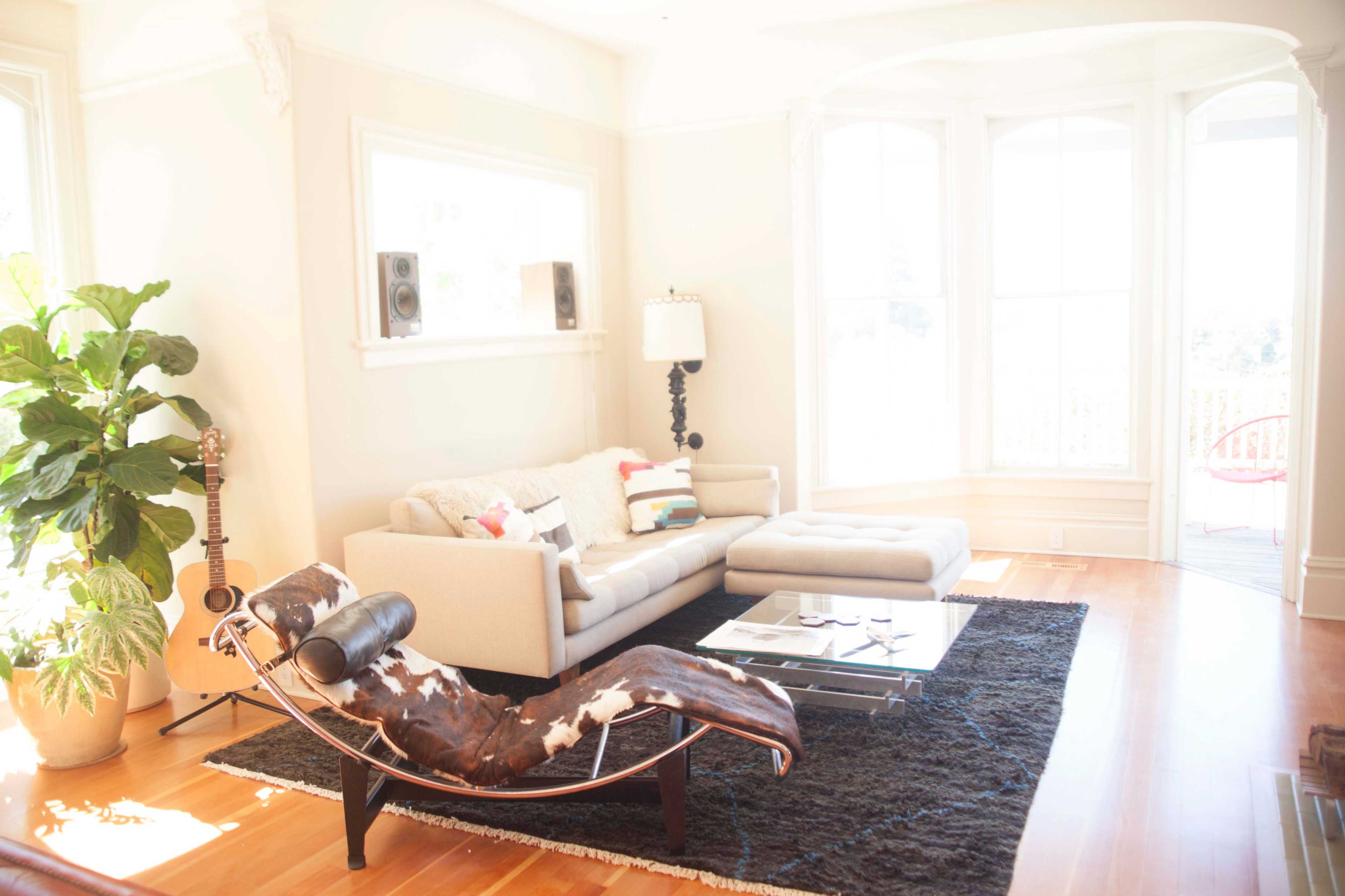 A bright living room features a light-colored couch, a large rug, a coffee table, and a modern chaise lounge, surrounded by windows and decorative plants.