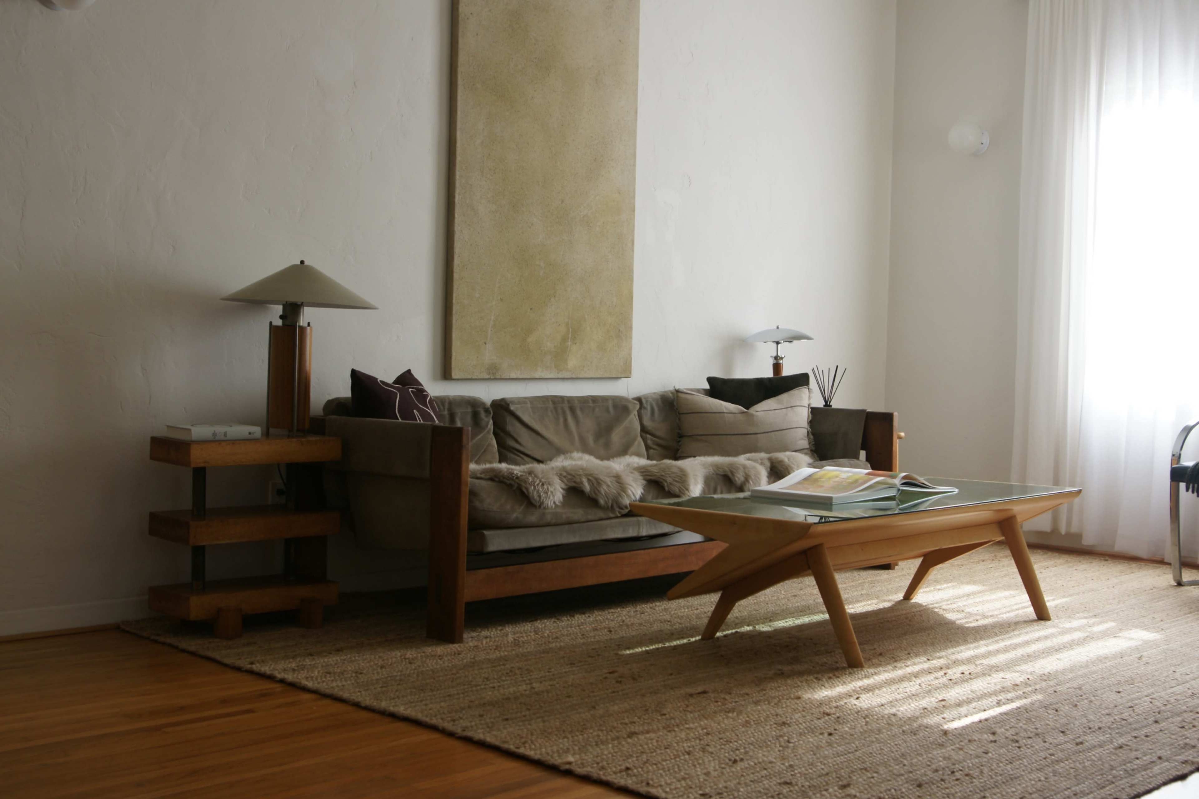 A modern living room features a wooden sofa with cushions, a glass-top coffee table, a textured rug, and a floor lamp, set against a light-colored wall with abstract art.