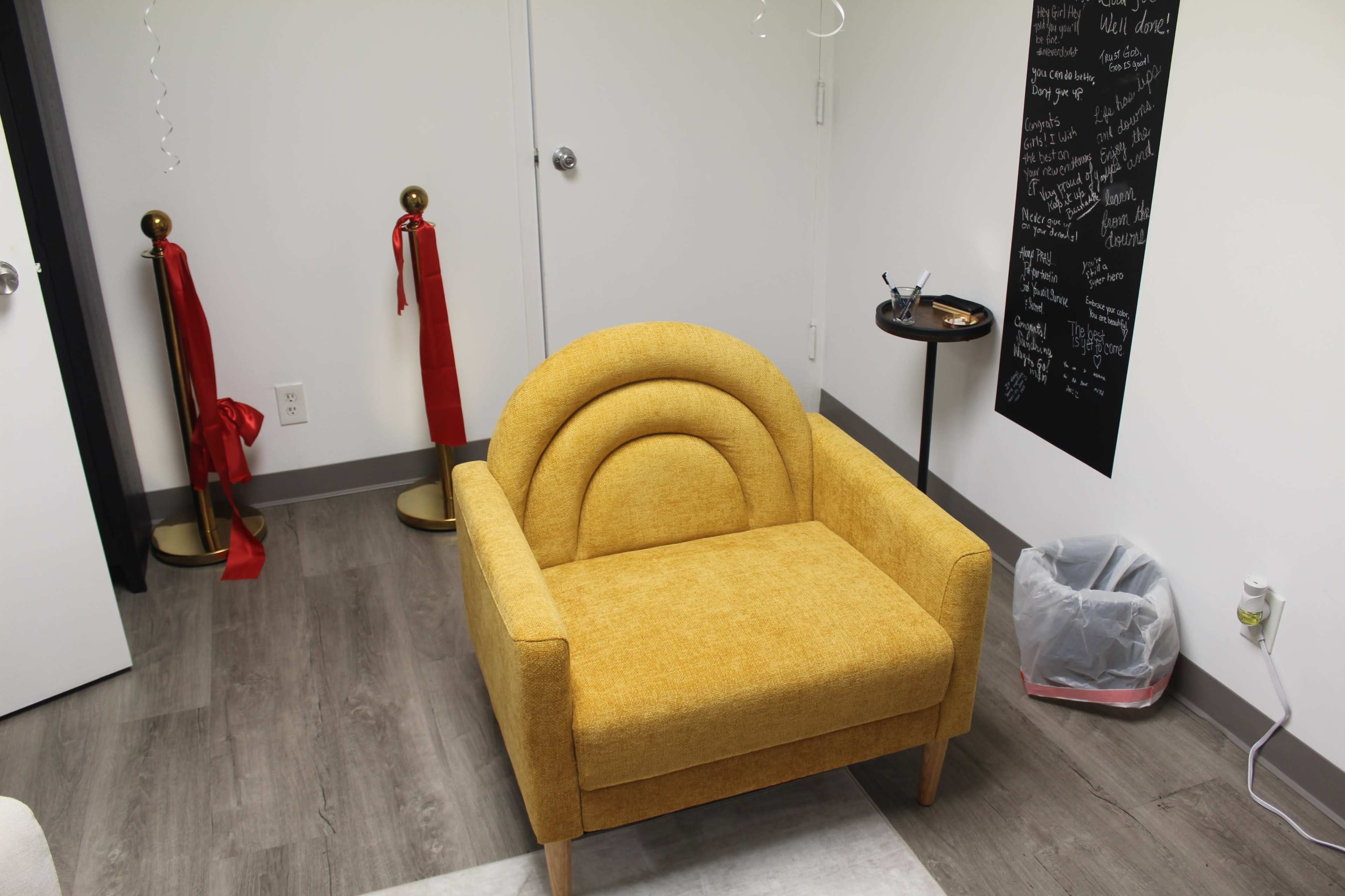 The image shows a yellow armchair with a distinctive curved backrest, positioned in a room with a wastebasket and a small round table nearby.