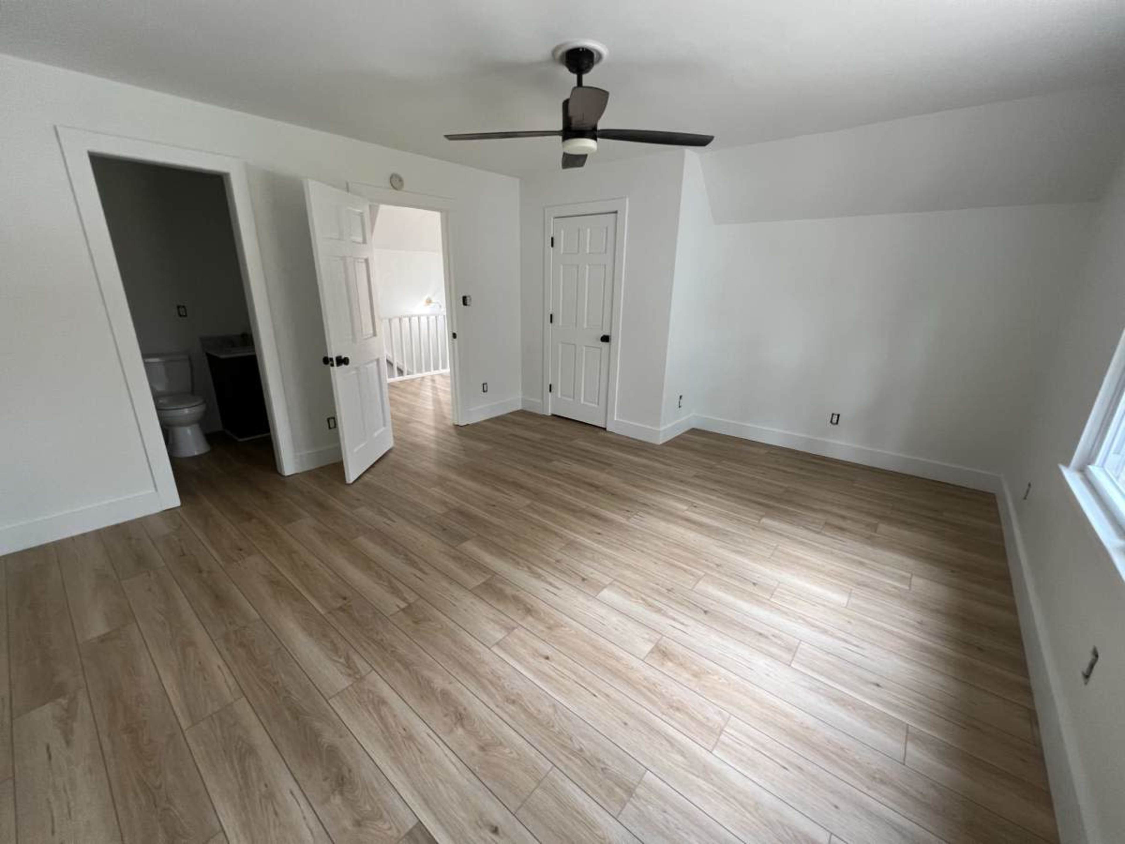 An empty room with light wood flooring, a ceiling fan, and two open doorways leading to what appears to be a bathroom and a hallway.