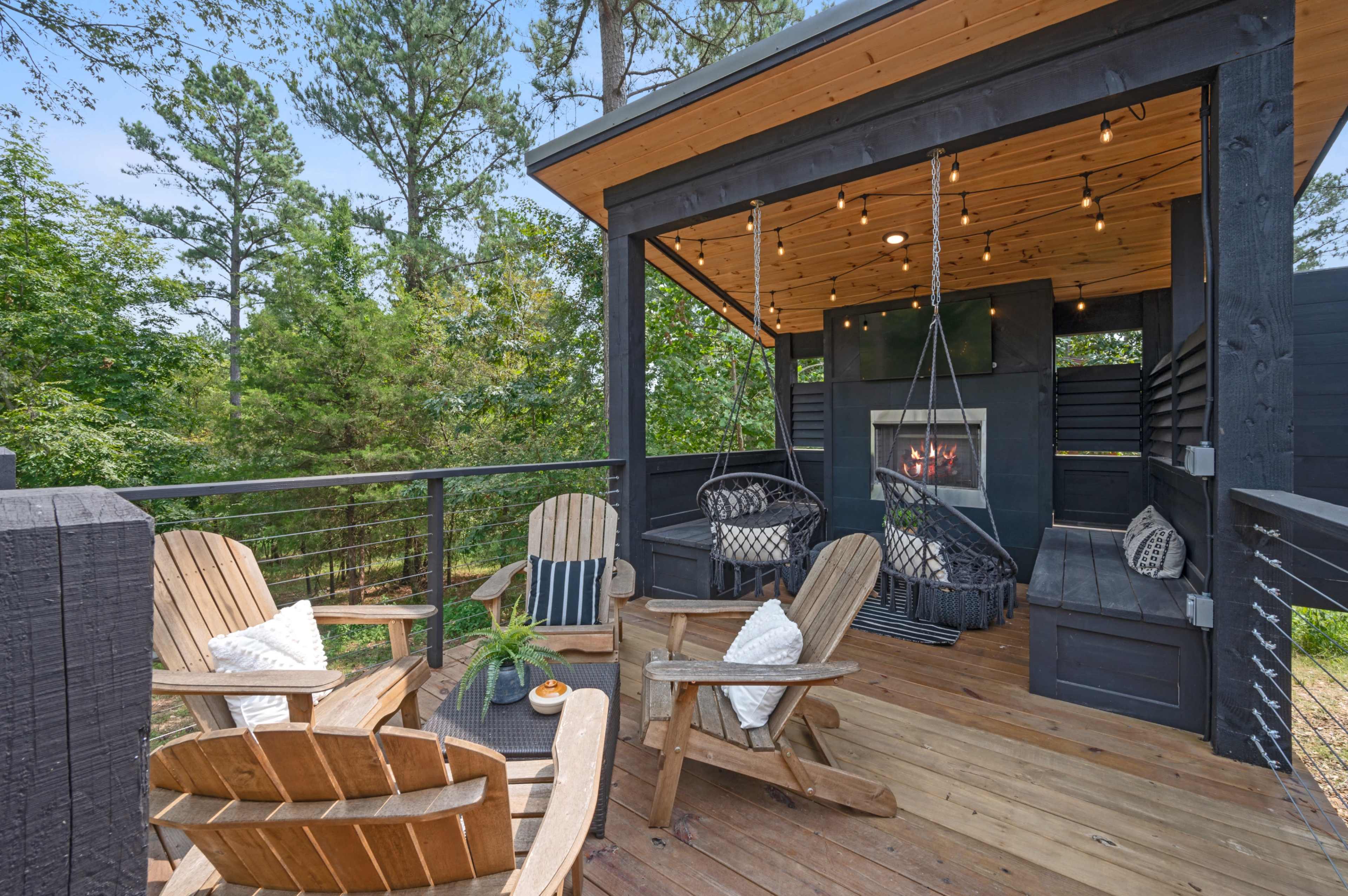 A wooden deck with several Adirondack chairs, a hanging chair, and a fireplace, surrounded by trees.