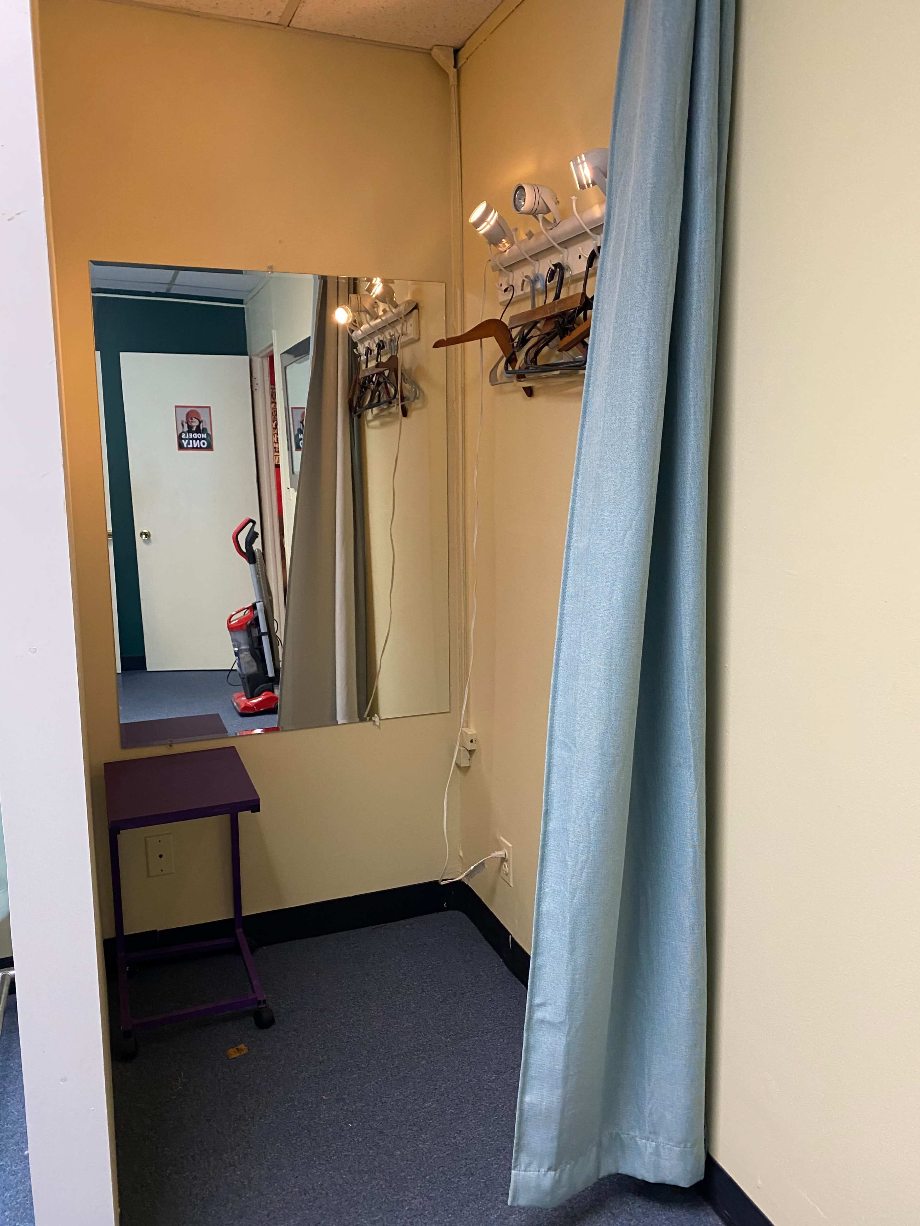 The image shows a small dressing area featuring a mirror, a blue curtain, a purple table, and several clothes hangers on the wall.