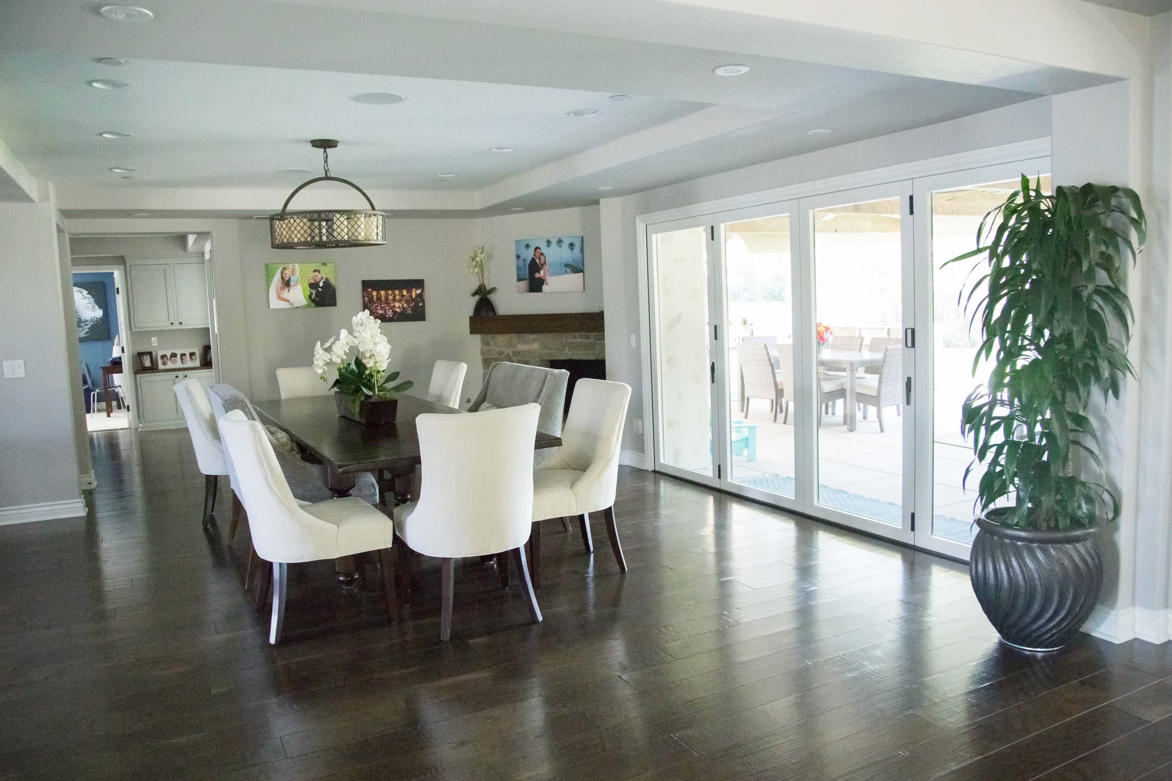 A modern dining room features a large wooden table surrounded by white chairs, with sliding glass doors leading to an outdoor seating area.
