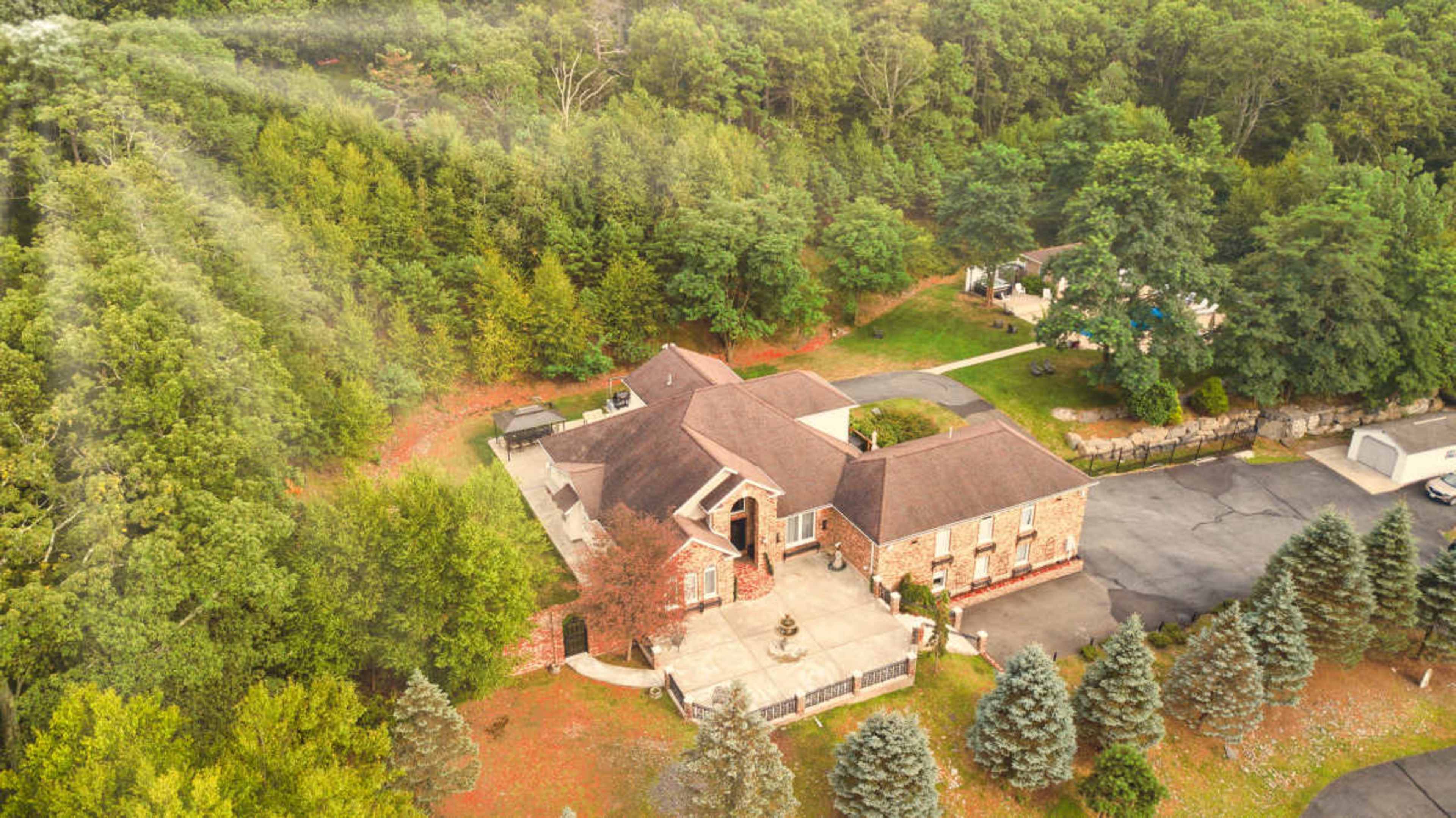 The image shows an aerial view of a large brick house surrounded by dense, green trees with a paved driveway and beautifully landscaped grounds.