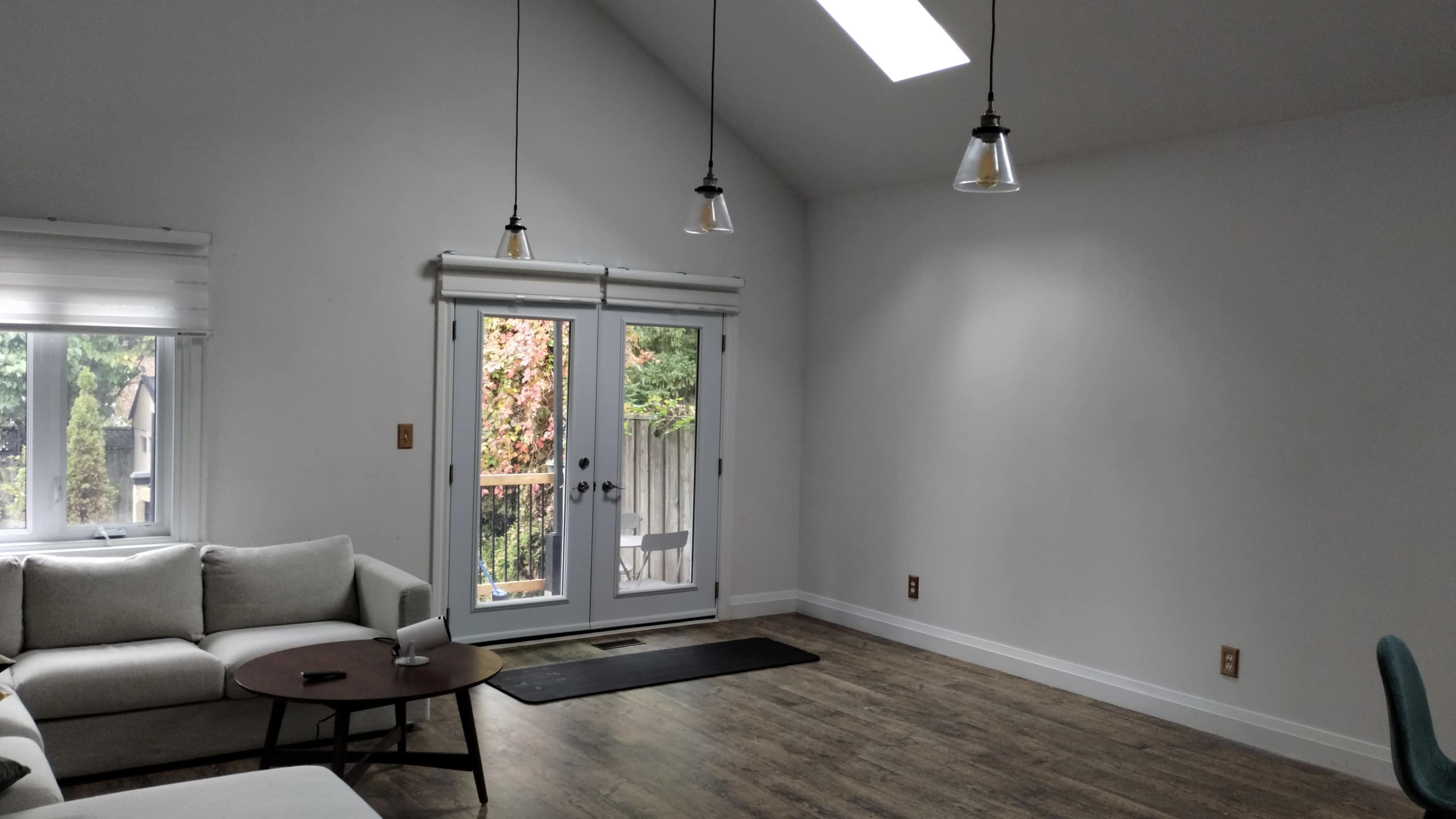 The living room features a light-colored sofa, a wooden coffee table, and a set of three pendant lights hanging from the sloped ceiling, with large glass doors leading to an outdoor deck.